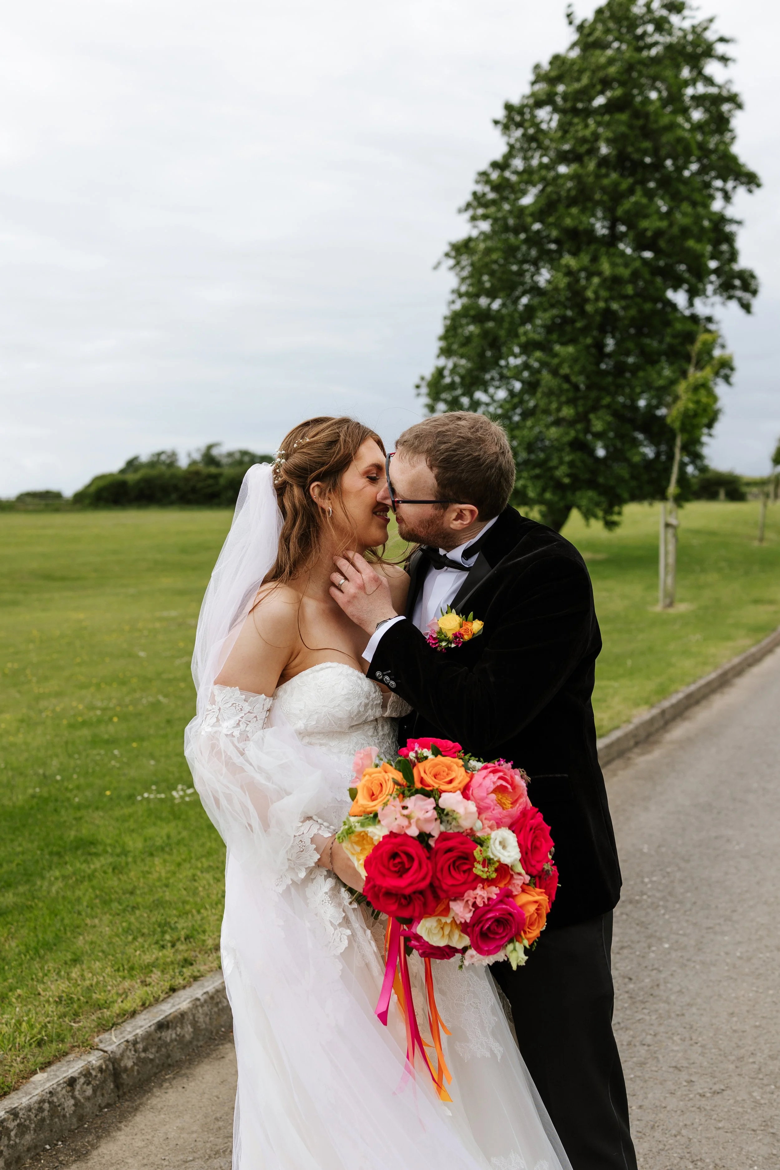 A bride and groom sharing a kiss outdoors, with the bride holding a colorful bouquet of flowers, in a grassy park with a tree and cloudy sky in the background.
