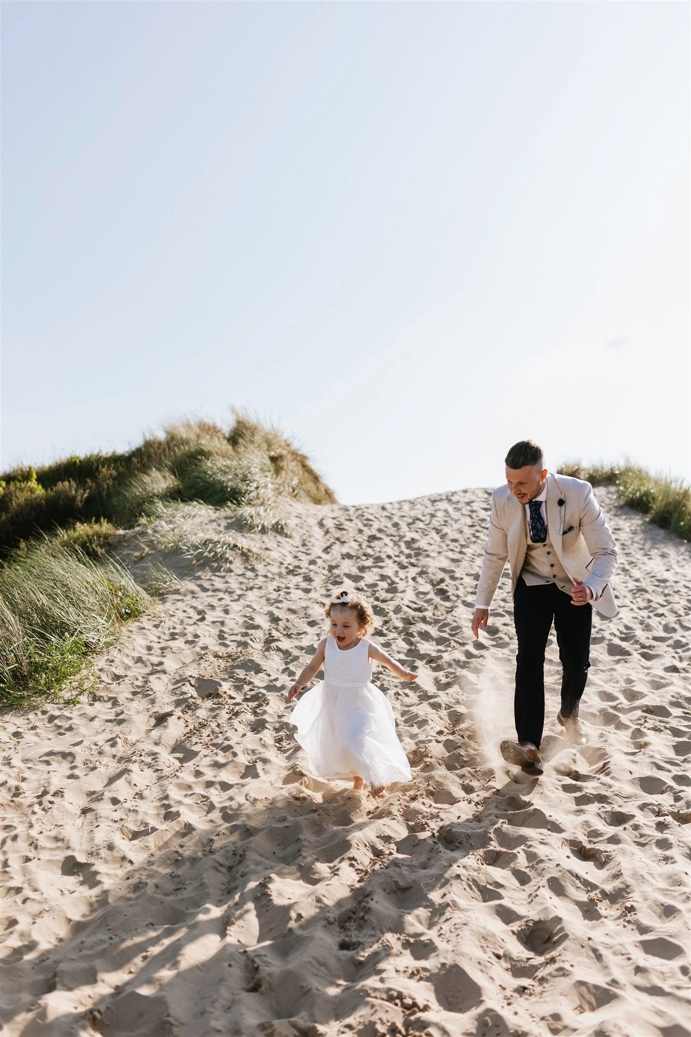 A man and a young girl running on a sandy beach with grass-covered dunes in the background under a clear sky.