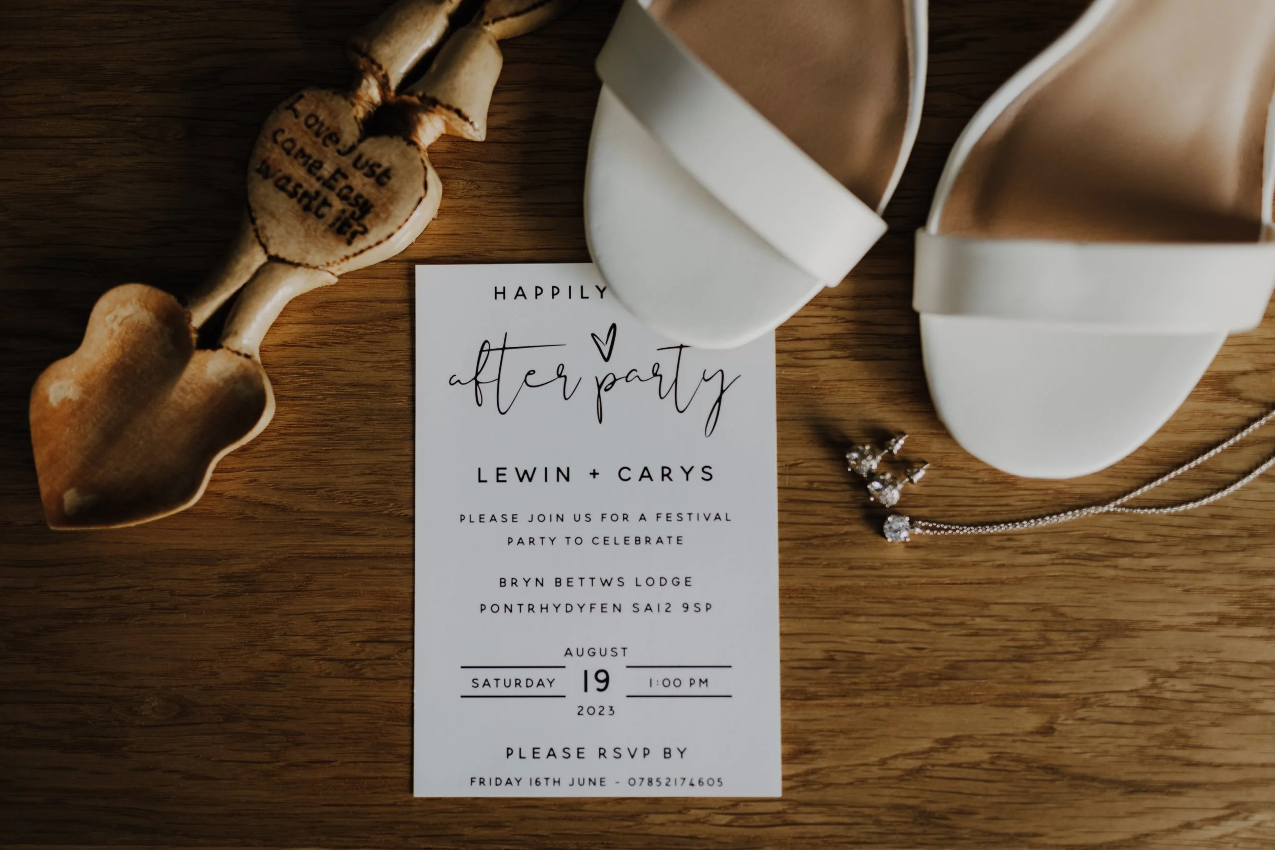 A wedding invitation card with the text 'Happily After Party' and details of the event, placed on a wooden surface next to a pair of white shoes, a jewelry piece, and a decorative accessory shaped like a bone with the words 'I love the one I came eas