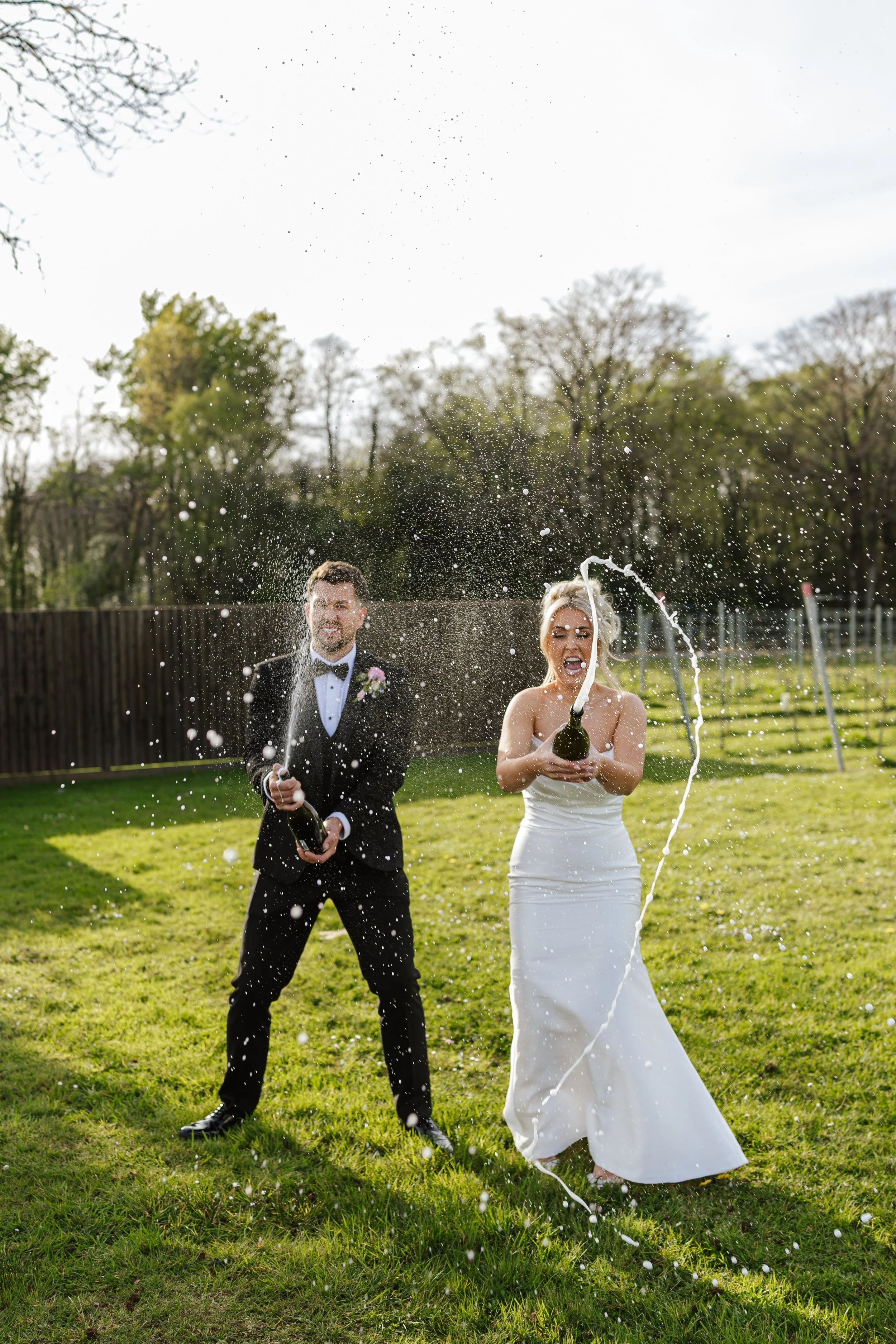 Bride and groom in wedding attire celebrating outdoors while opening champagne, with champagne spray and trees in the background.