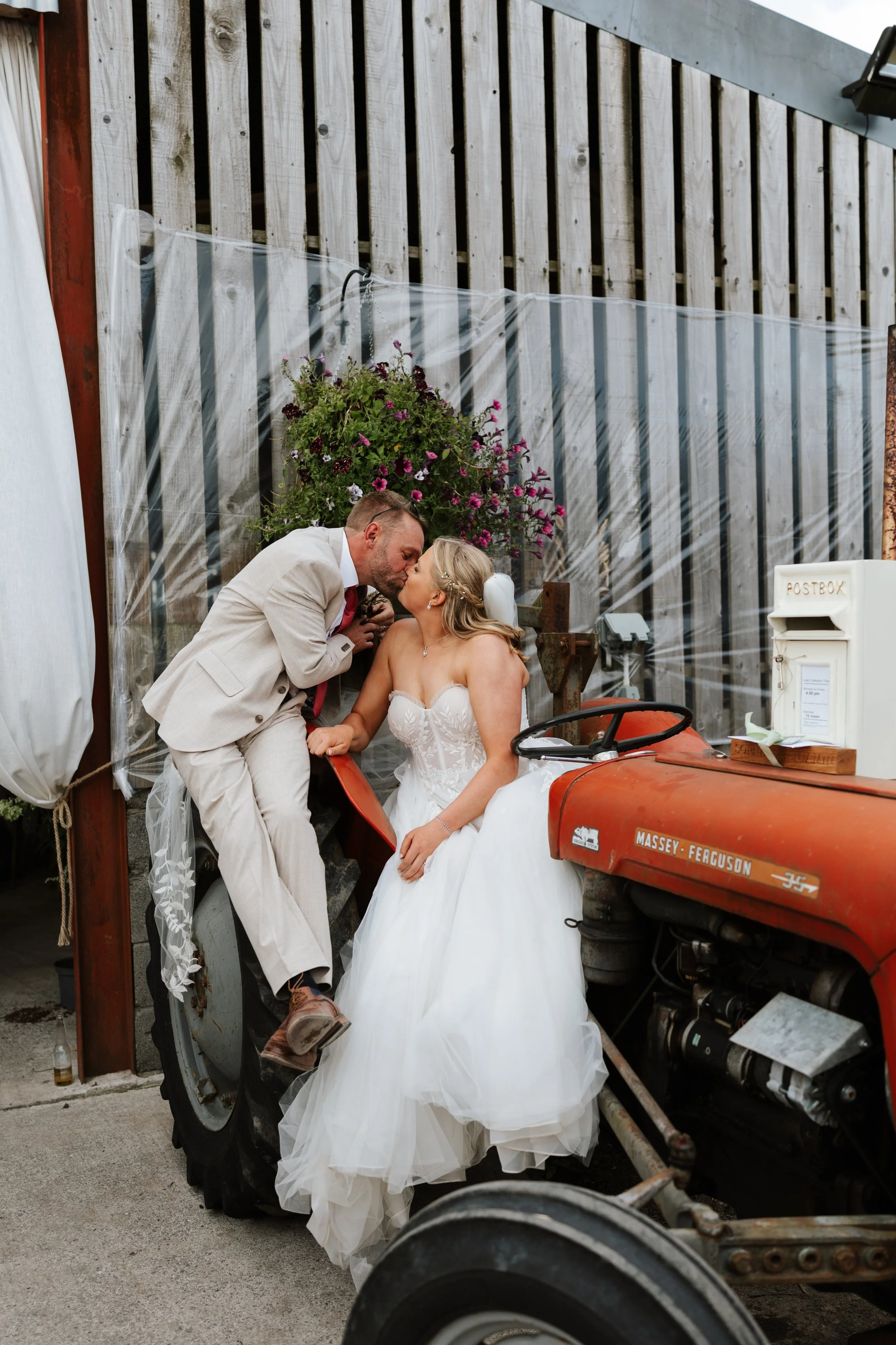 A bride and groom sharing a kiss on a vintage tractor, with a wooden fence and flowers in the background.