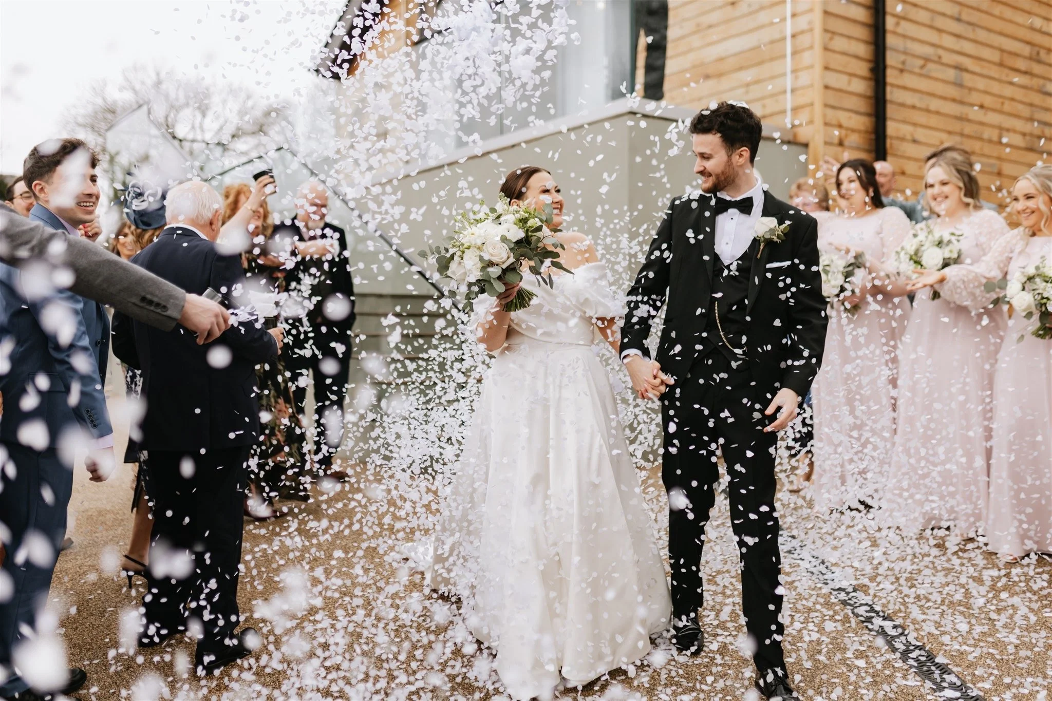 A bride and groom walking hand in hand amid confetti during their wedding celebration, surrounded by guests outdoors.