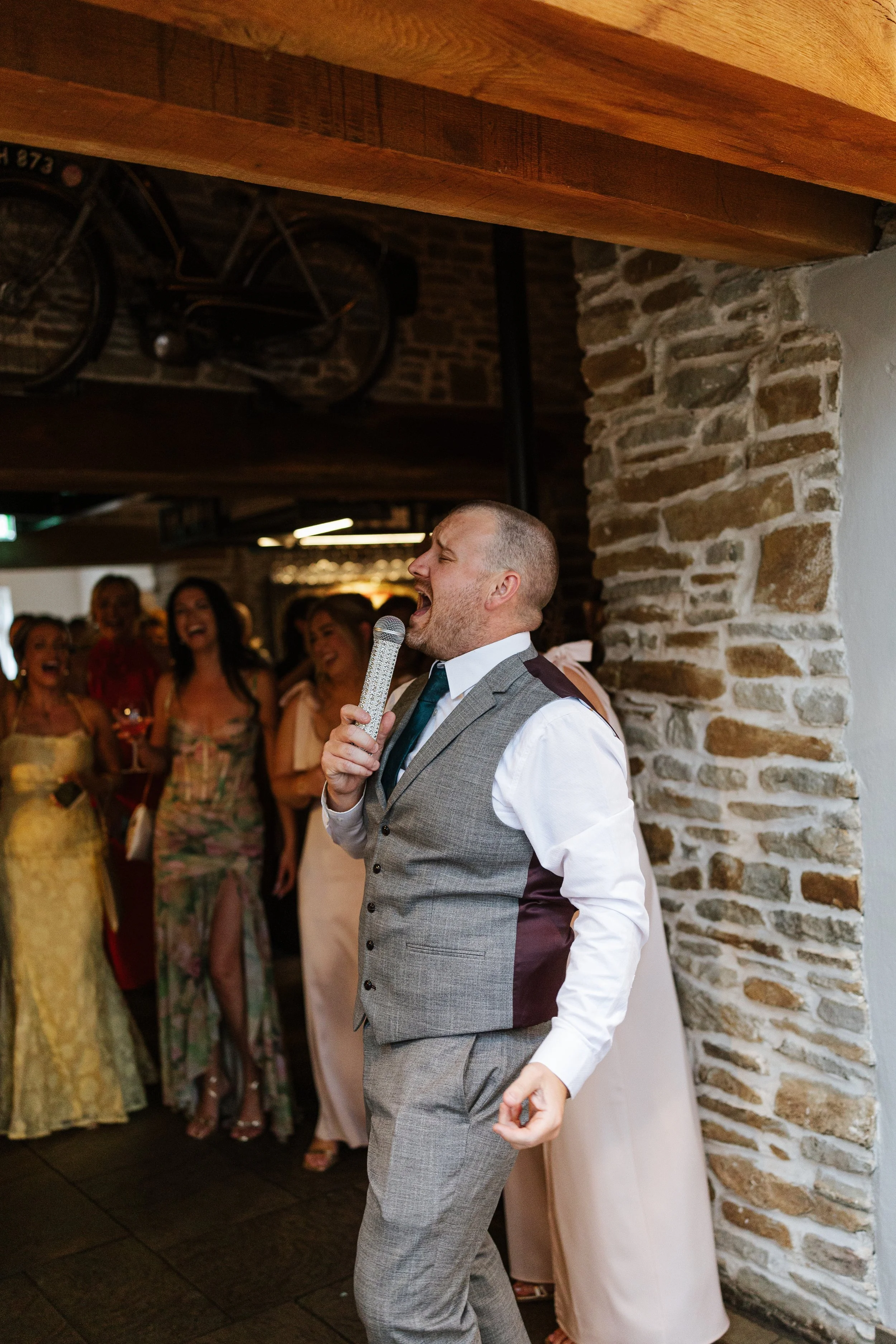 A man in a gray vest and white shirt singing into a microphone at a wedding reception, surrounded by guests in colorful dresses, in a rustic venue with exposed brick walls.