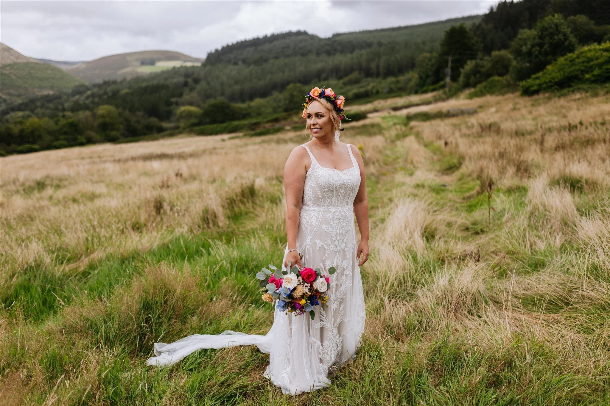 A woman in a wedding dress holding a bouquet of flowers standing in a grassy field with rolling hills and trees in the background.