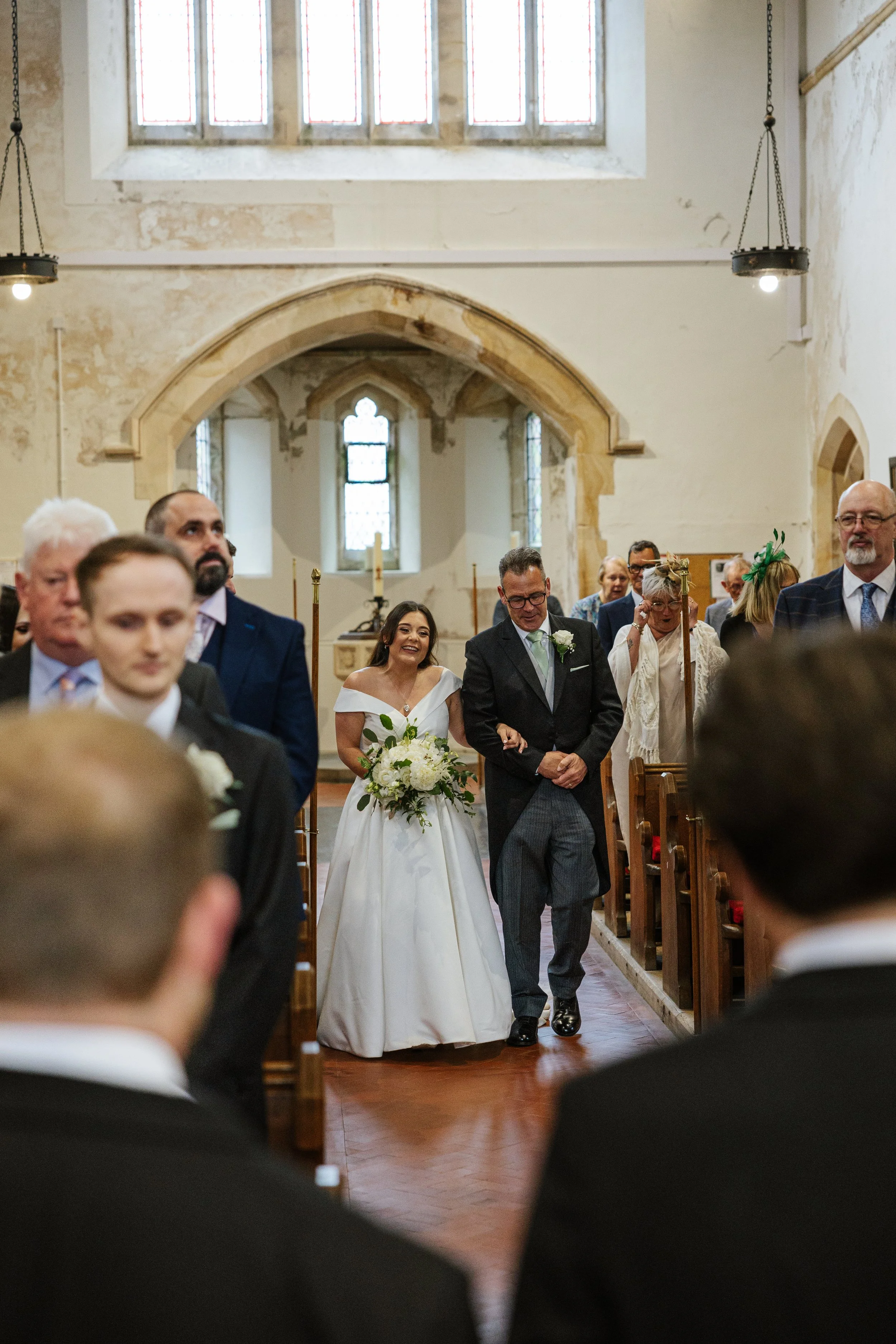 A bride in a white wedding gown walking down the aisle of a church with a man in a gray suit, likely her father. The church has high ceilings, stained glass windows, and several guests seated in the pews, some smiling and watching the bride.