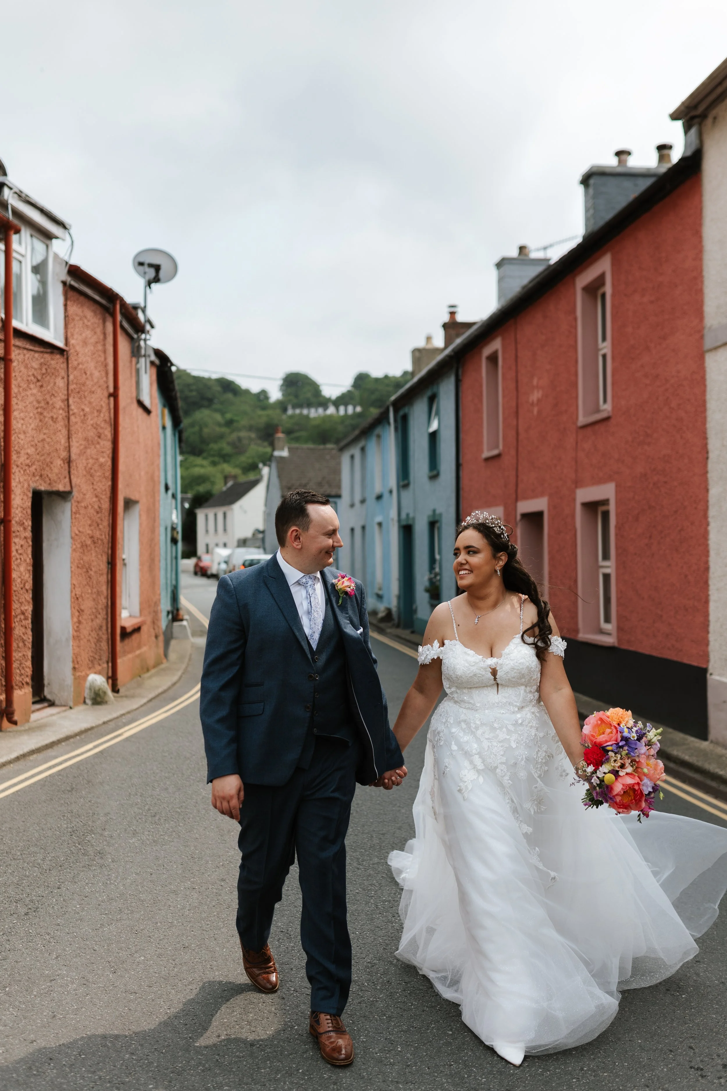 A bride and groom walking hand in hand down a colorful street, smiling at each other. The bride is wearing a white wedding gown and holding a large bouquet of colorful flowers. The groom is dressed in a dark suit with a light-colored tie and boutonni