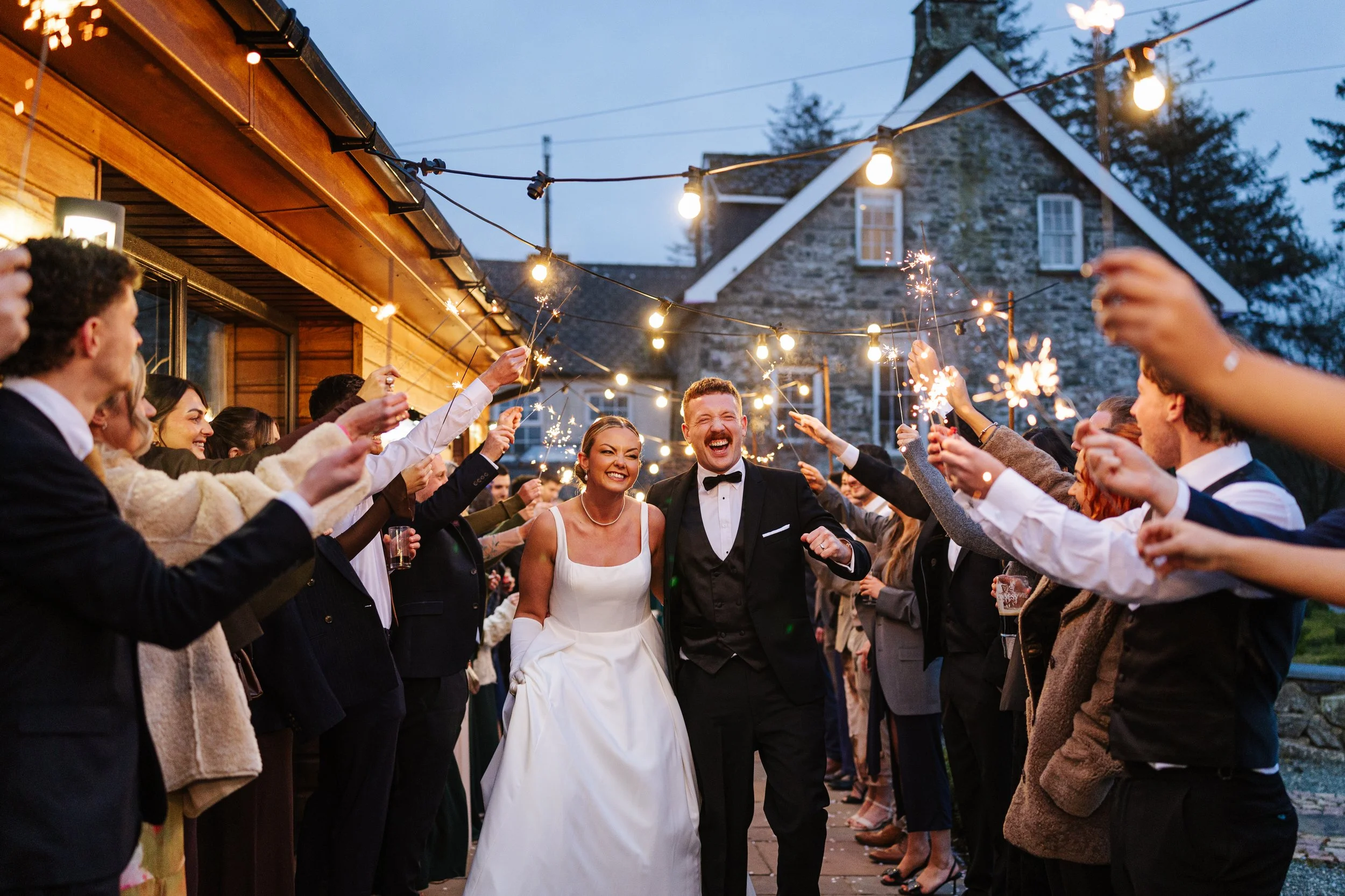 People at a wedding celebration outdoors during evening, holding sparklers, with a bride and groom in the center, smiling, under string lights, in front of a stone house.