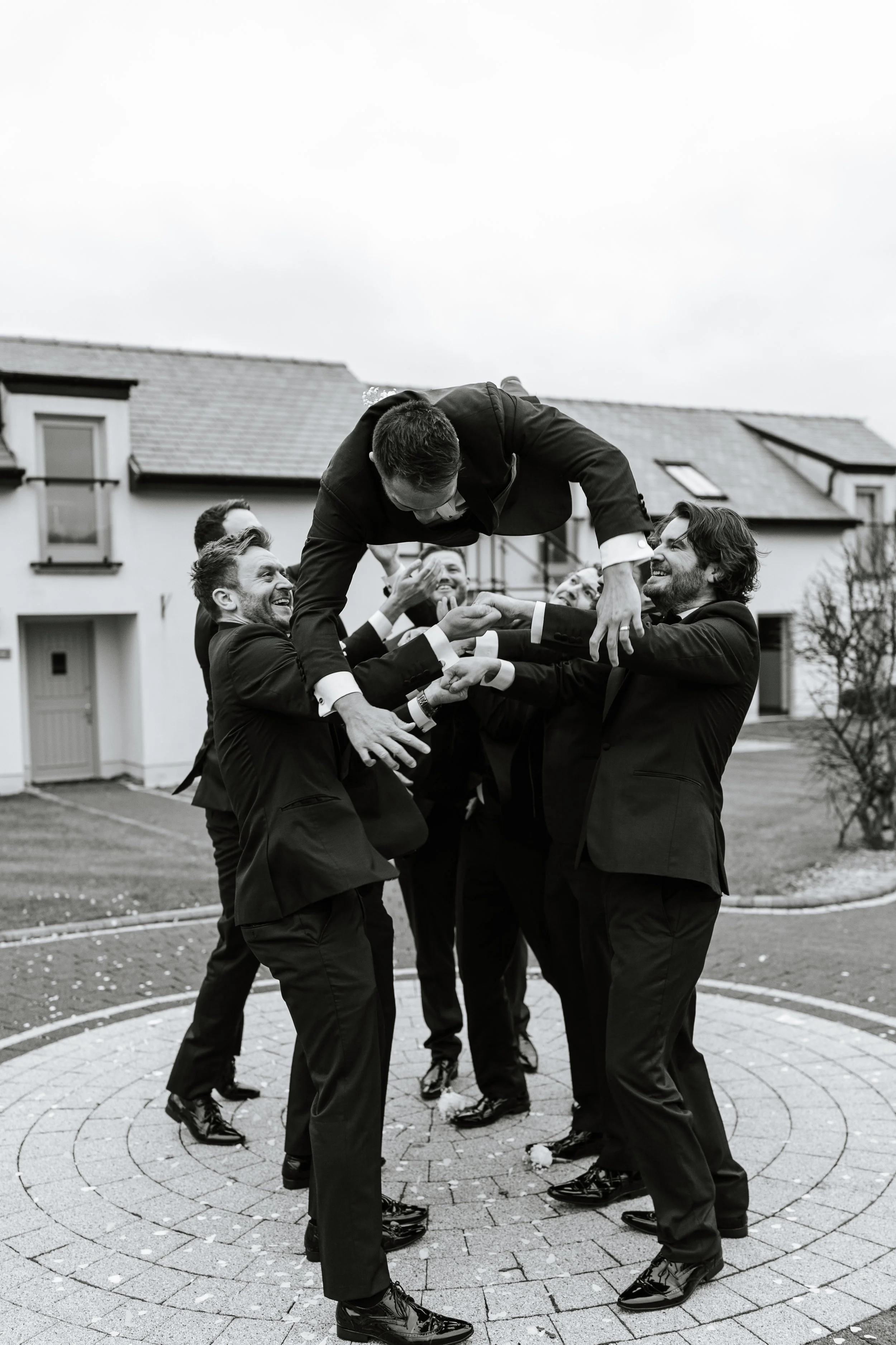 Group of men in tuxedos and dress shoes forming a circle outdoors, with one man being lifted by others, all smiling and laughing.