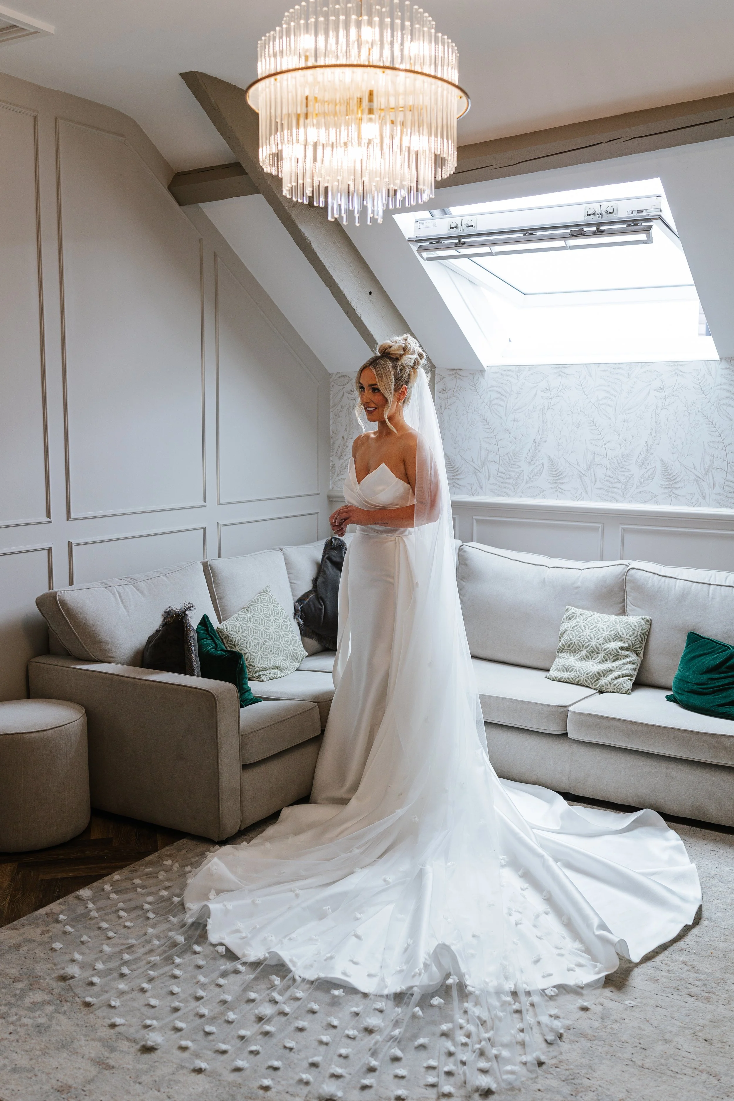 A bride in a strapless white wedding gown with a long train and veil standing in a bright, elegant living room with a large cream-colored sectional sofa, decorative pillows, a patterned rug, a modern chandelier, and a skylight with blinds.