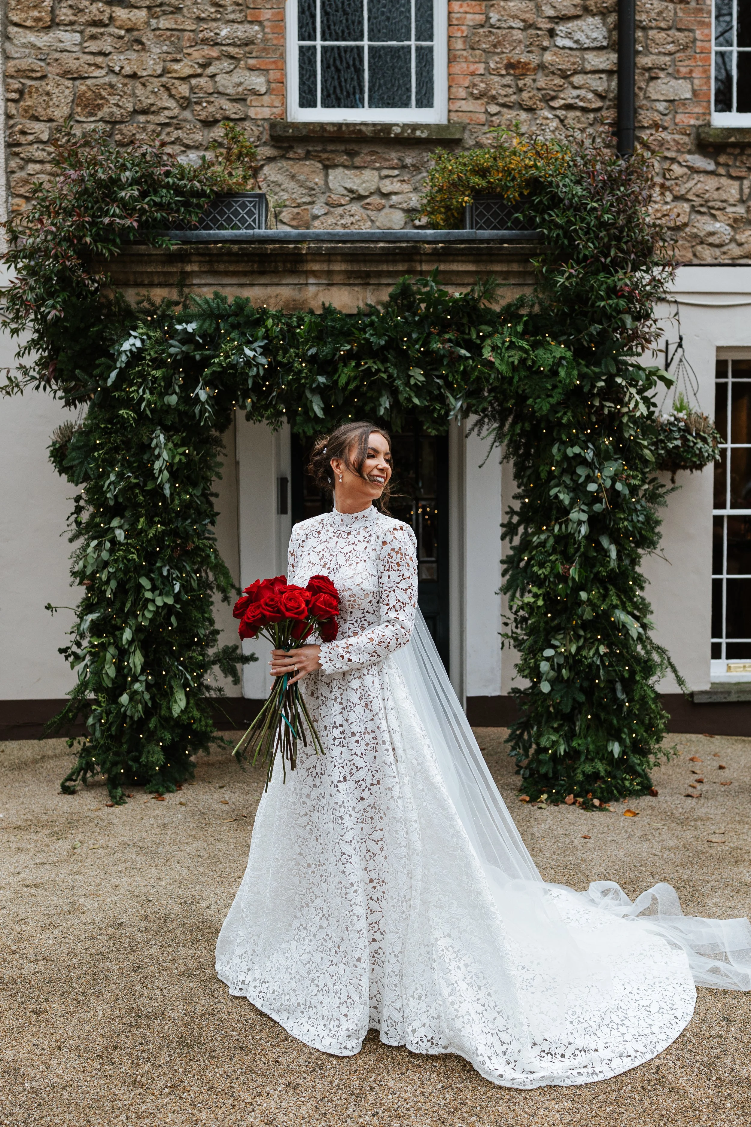 A smiling bride in a white lace wedding dress holding a bouquet of red roses standing in front of a decorated archway.