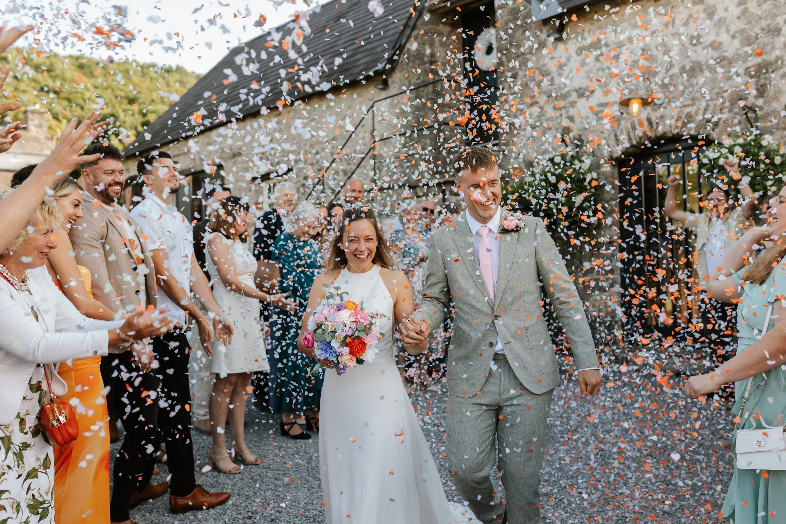 A newlywed couple walking hand-in-hand through a crowd of wedding guests, surrounded by falling colorful confetti during their outdoor celebration.