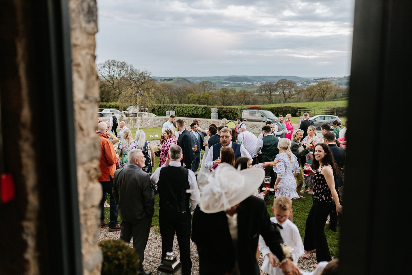 People attending a wedding reception outdoors during daytime, gathered on a lawn with scenic countryside in the background, some holding drinks and engaging in conversations.