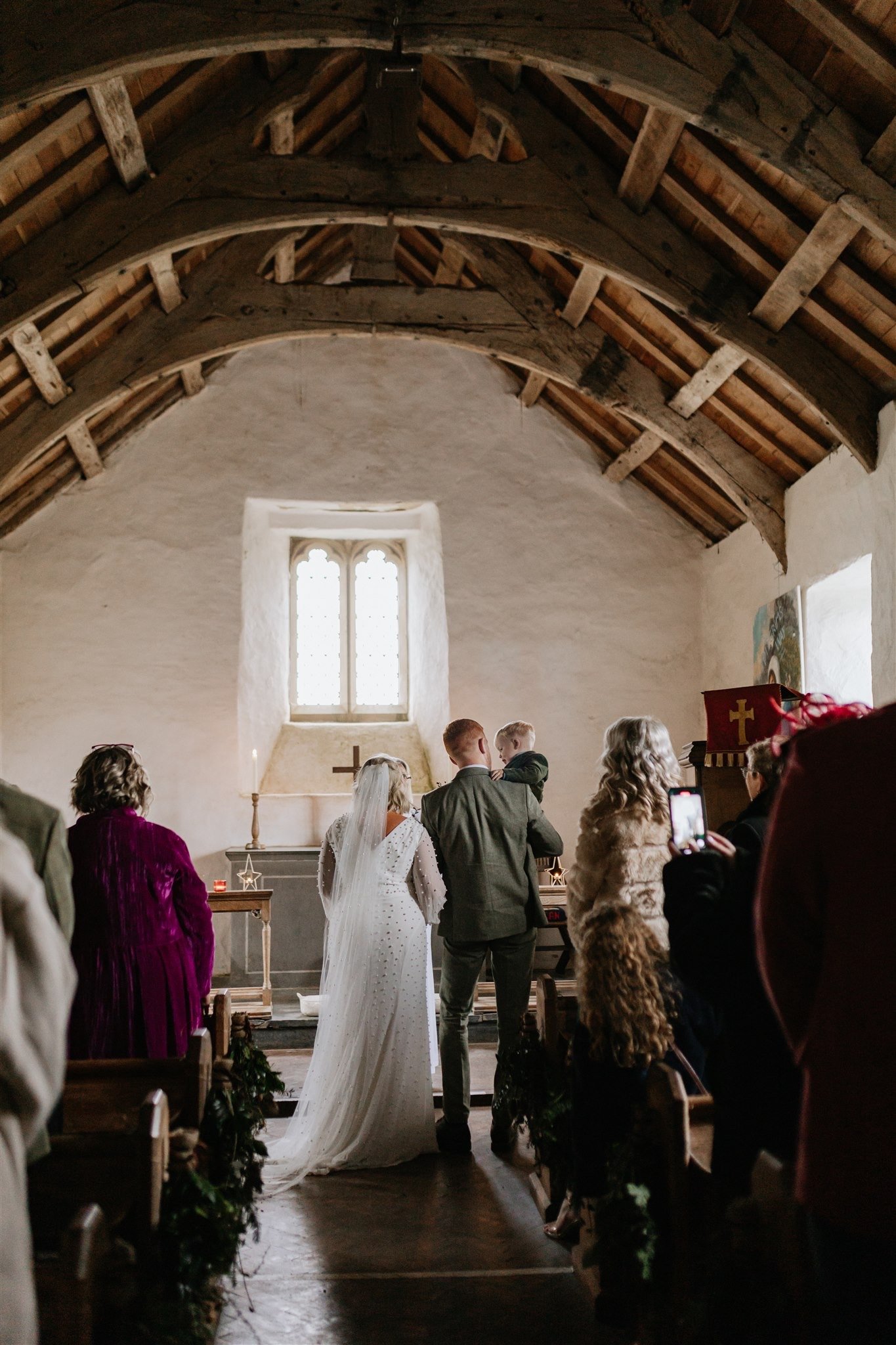 A wedding ceremony taking place inside a small, rustic chapel with wooden beams and a stained-glass window. The bride and groom stand at the altar, facing away from the camera, with guests seated on either side. The bride is wearing a white veil and 