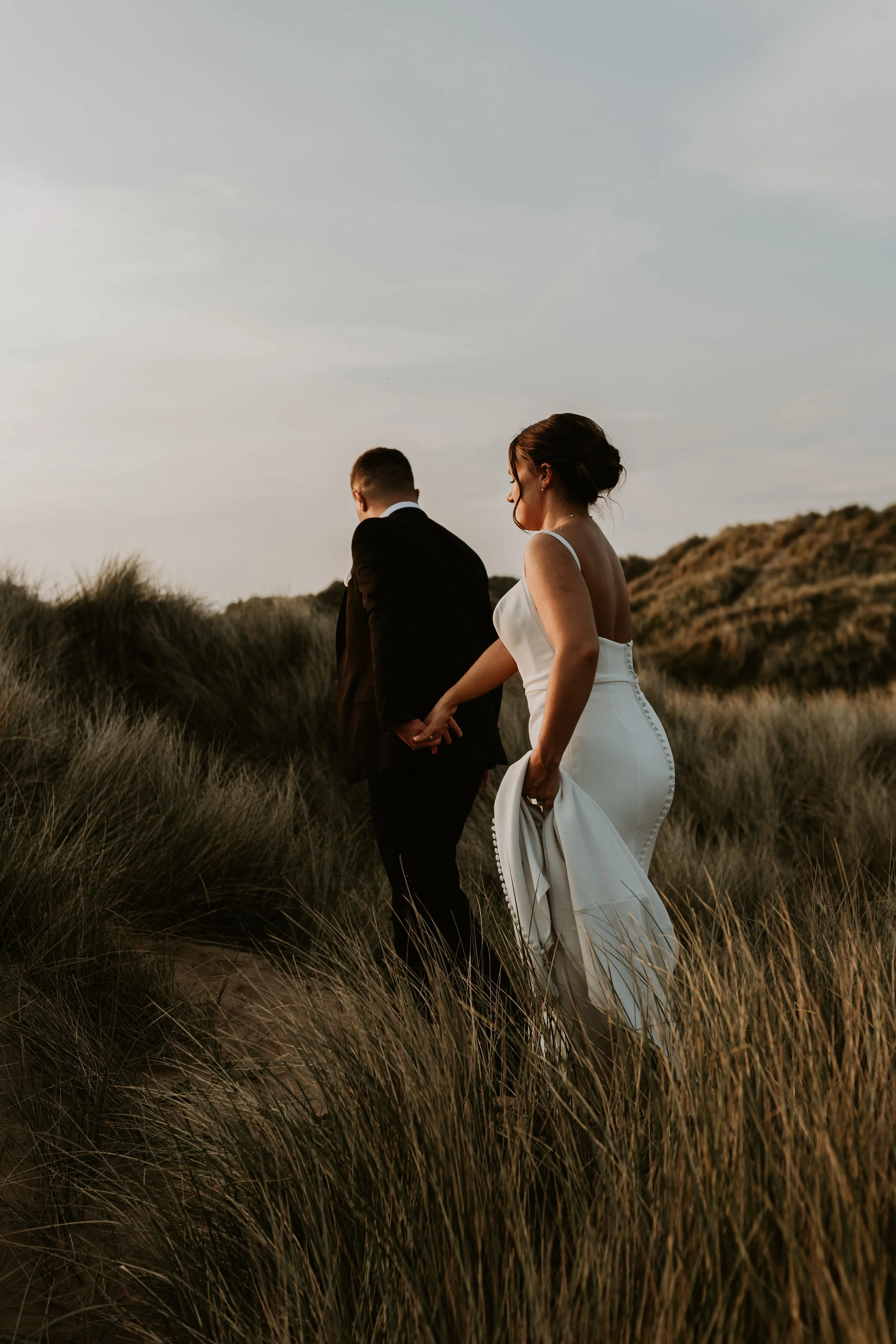 A bride and groom holding hands and walking through a sandy, grassy landscape at sunset
