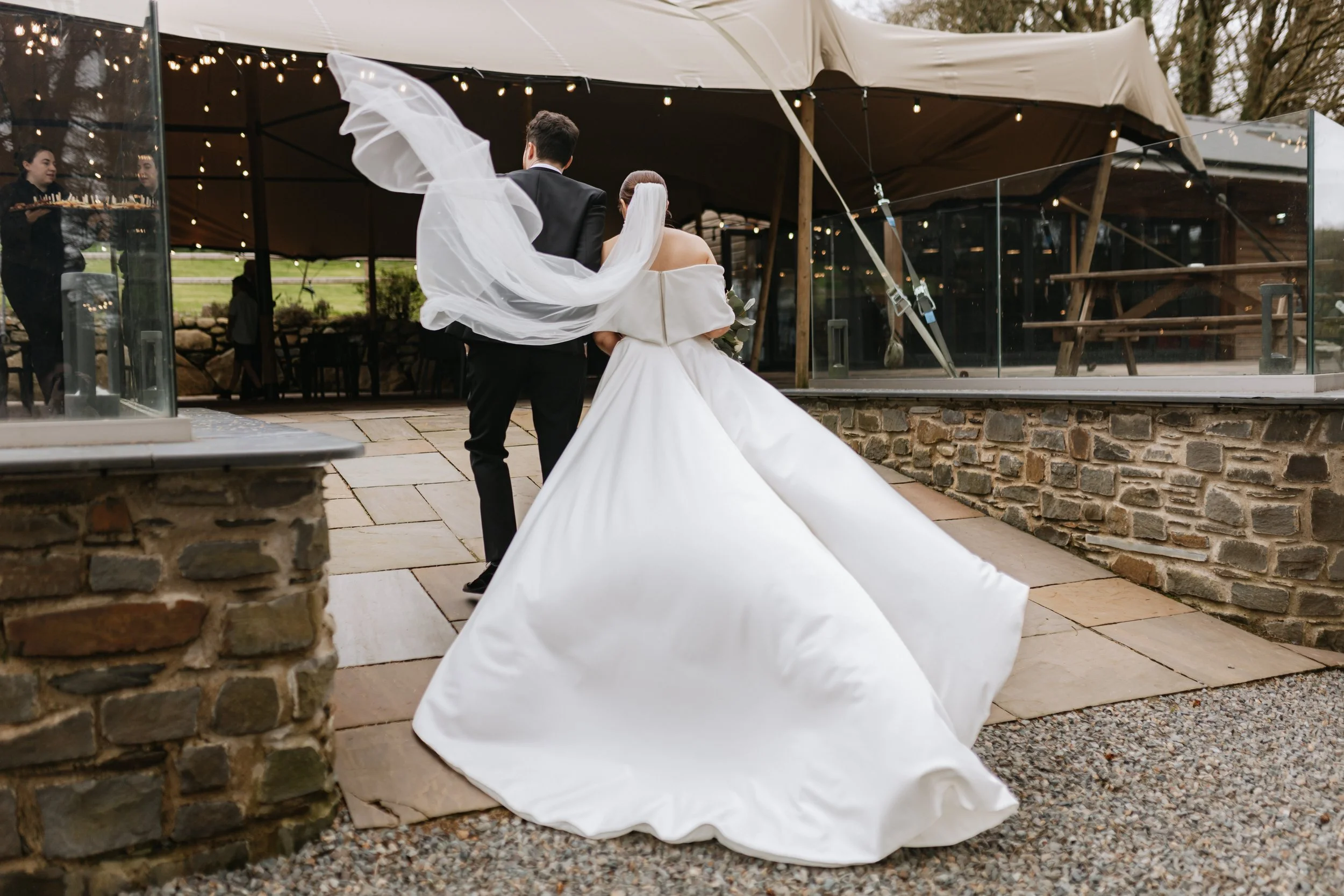 Bride and groom standing together during outdoor wedding ceremony, with bride holding flowers and groom dressed in black tuxedo, under a canopy decorated with string lights.