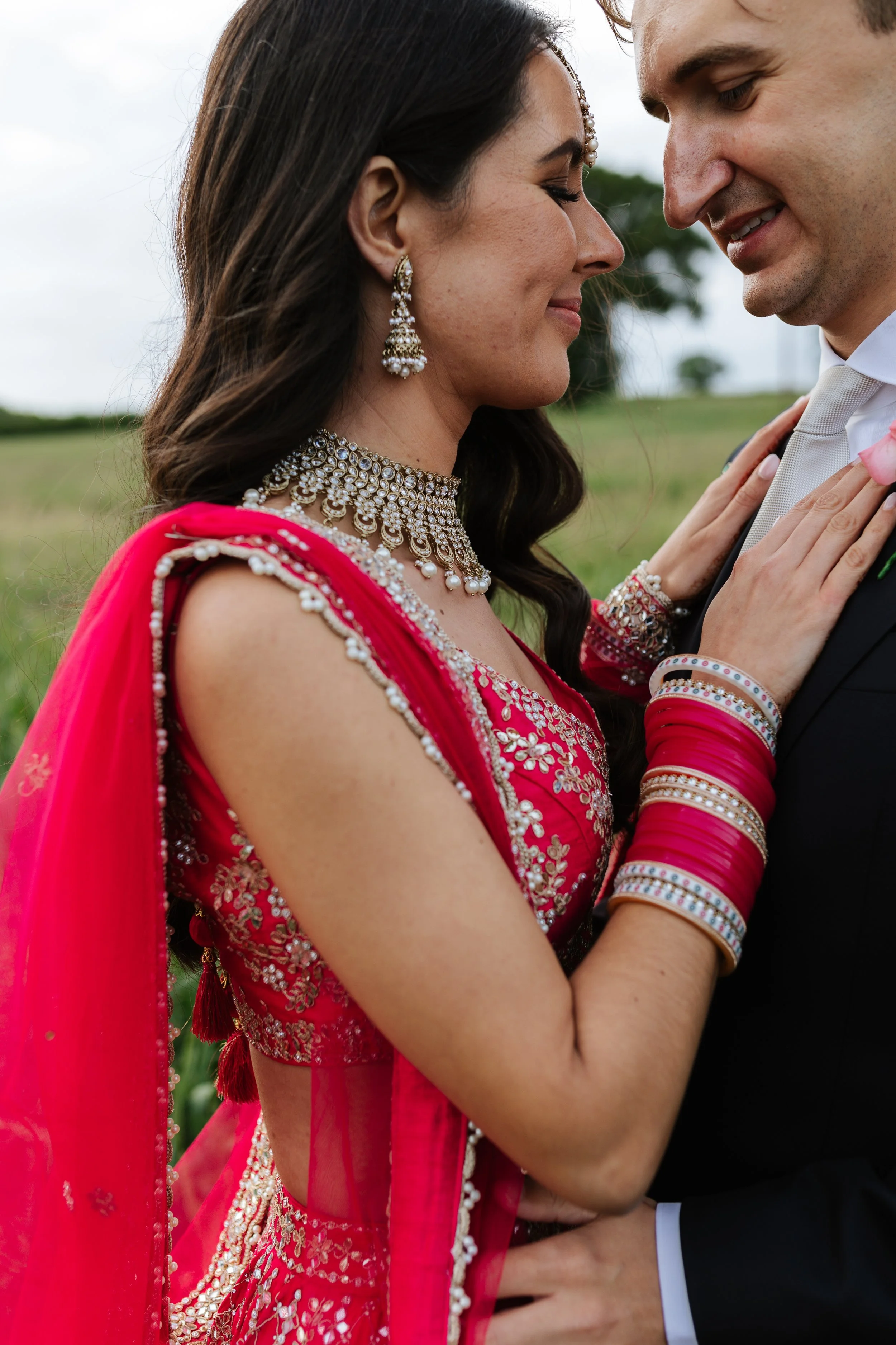 A couple in traditional Indian attire closely holding each other, with their foreheads touching and eyes closed, outdoors in a green field.