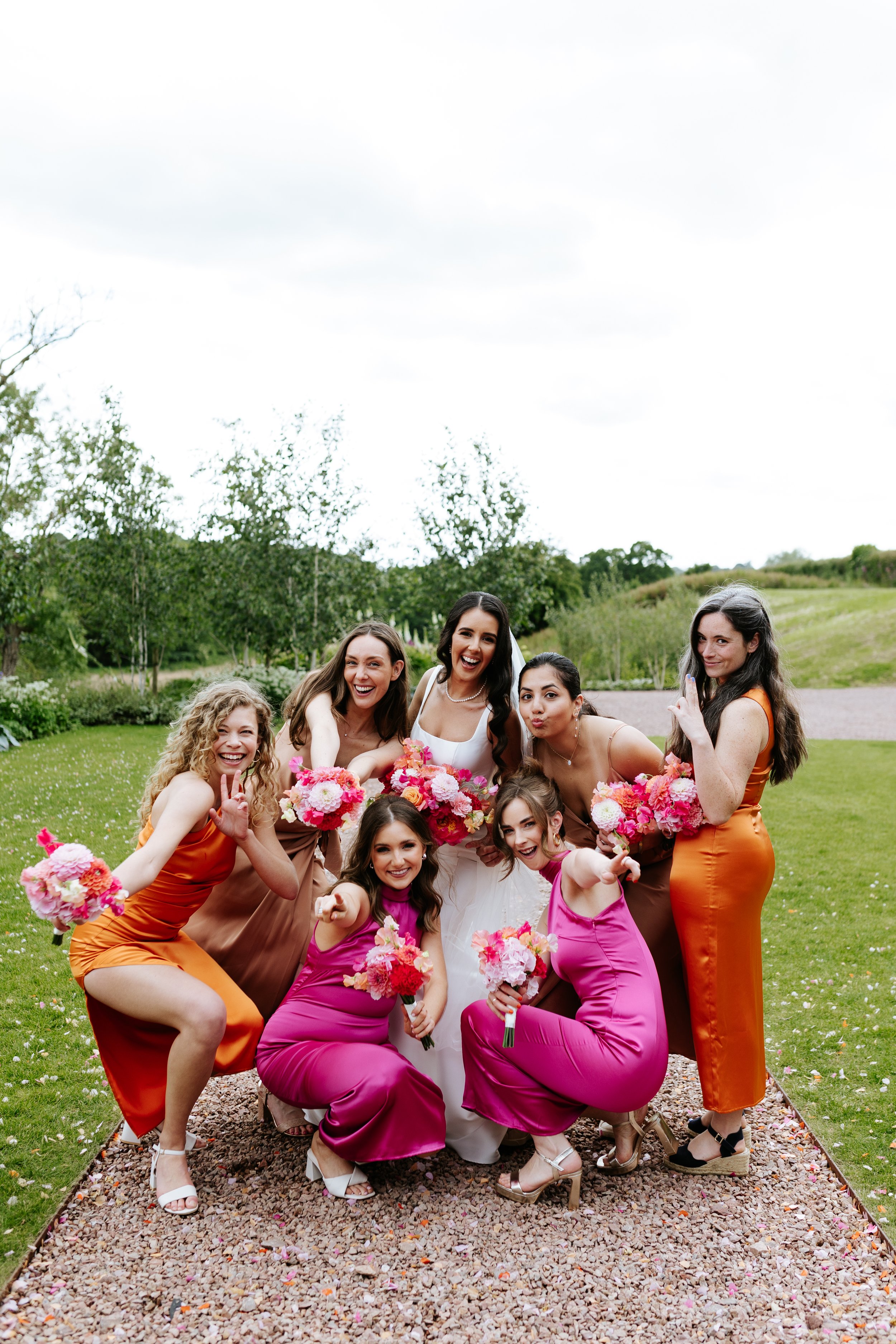 Group of women, including a bride in white, holding pink and red flower bouquets, posing outdoors on a gravel pathway with greenery and trees in the background, celebrating a wedding.