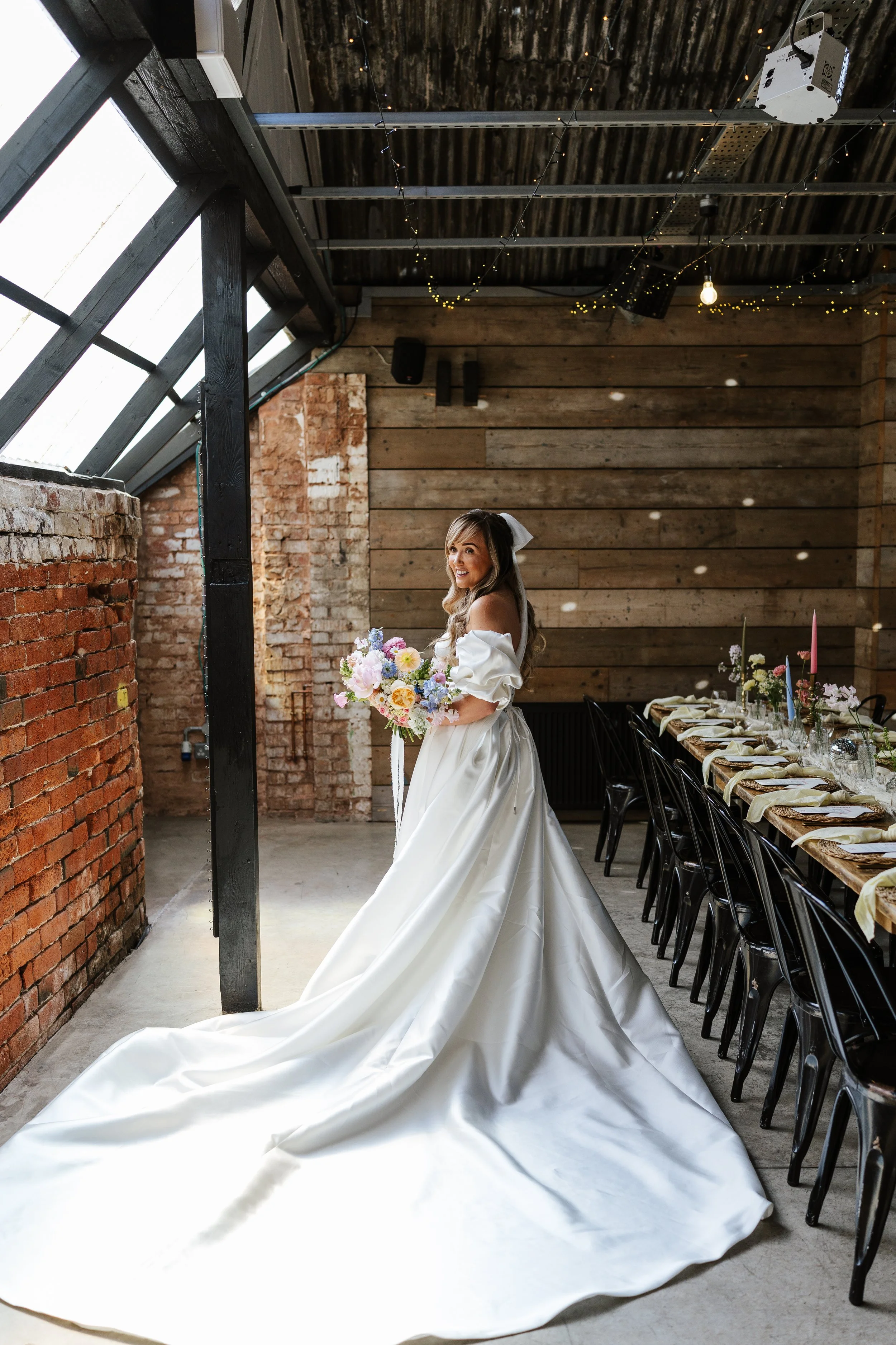 A bride in a white wedding gown holding a bouquet standing in a rustic event space with brick and wooden walls, decorated with string lights, near a long table set for a celebration.