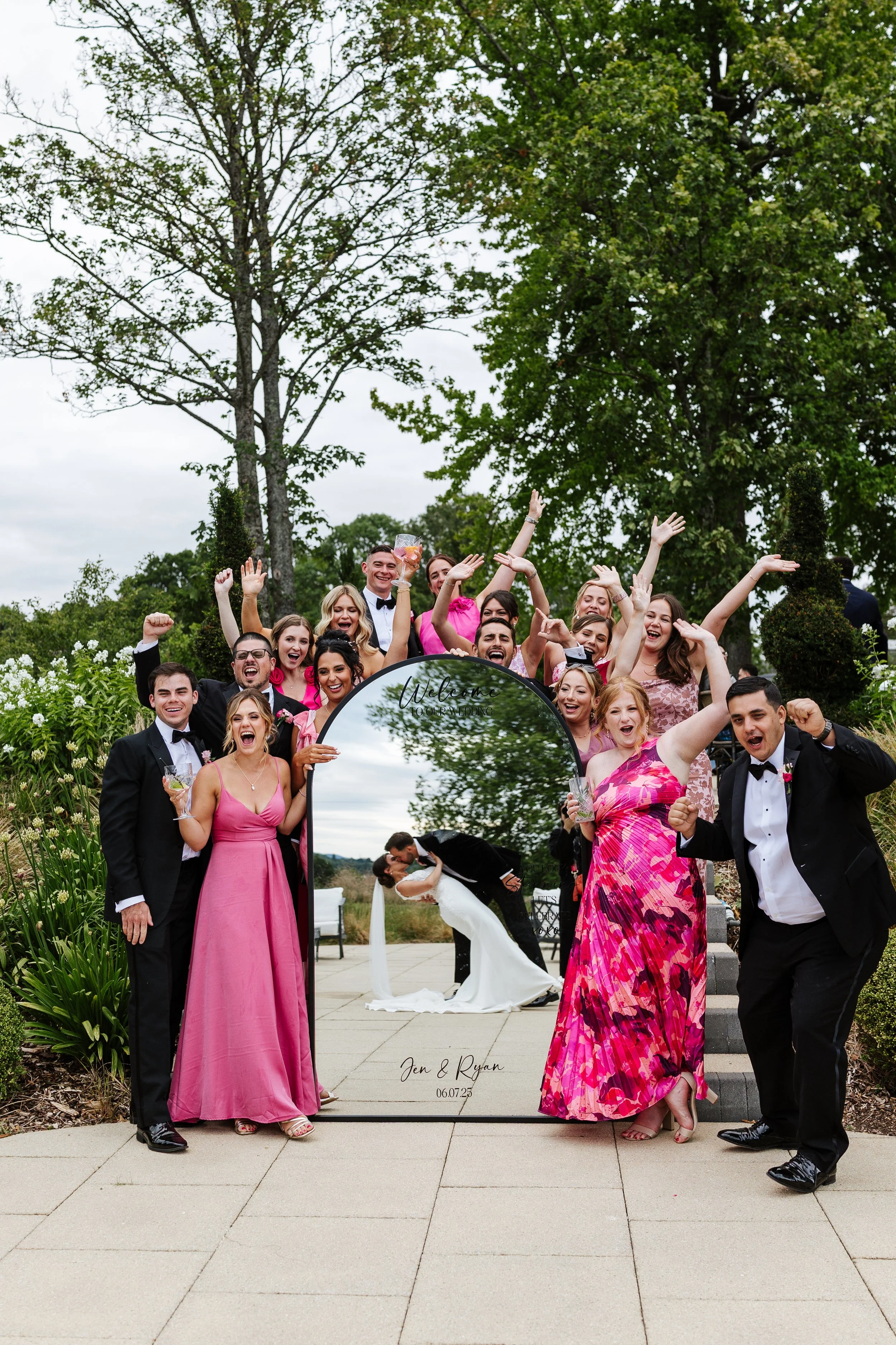 Group of people celebrating at a wedding outside, with a couple kissing in the background. A mirror with a sign reads 'Welcome to our wedding' and the date '06.07.23'.