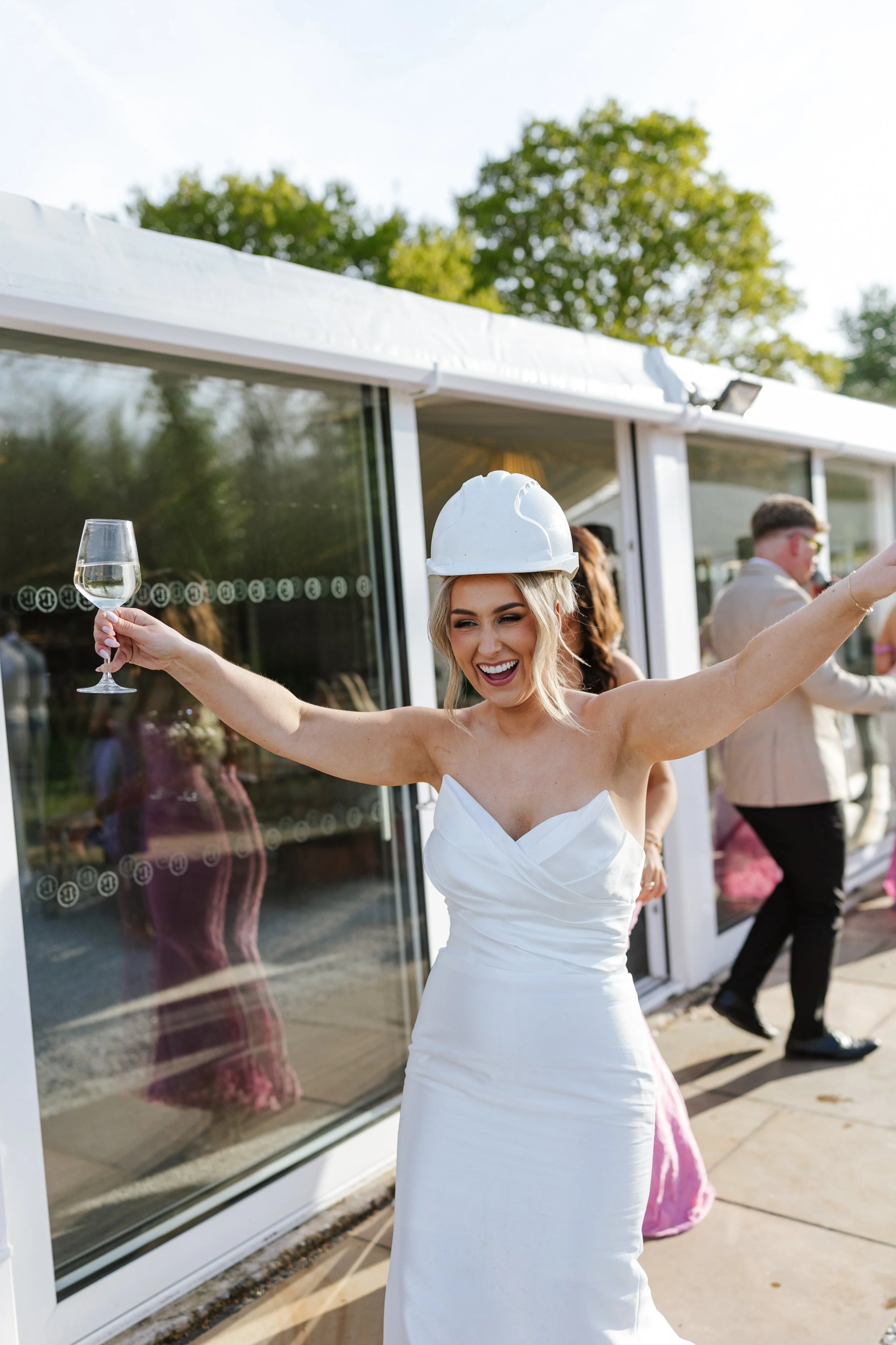 A woman in a white dress and white hard hat is holding a glass of white wine and smiling at an outdoor event.