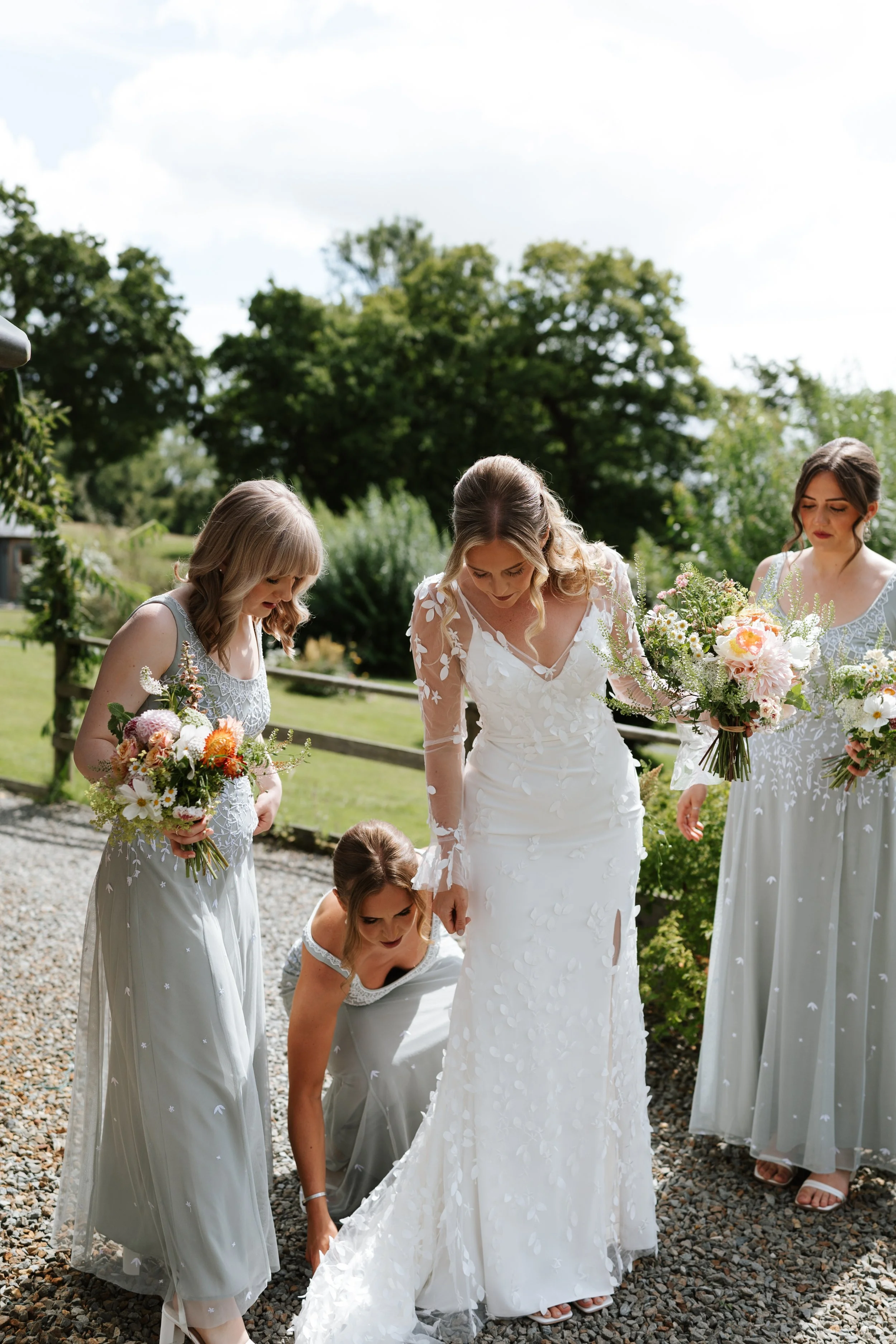 A bride in a white wedding dress surrounded by four bridesmaids in light gray dresses with floral bouquets, outdoors on a sunny day with green trees in the background.