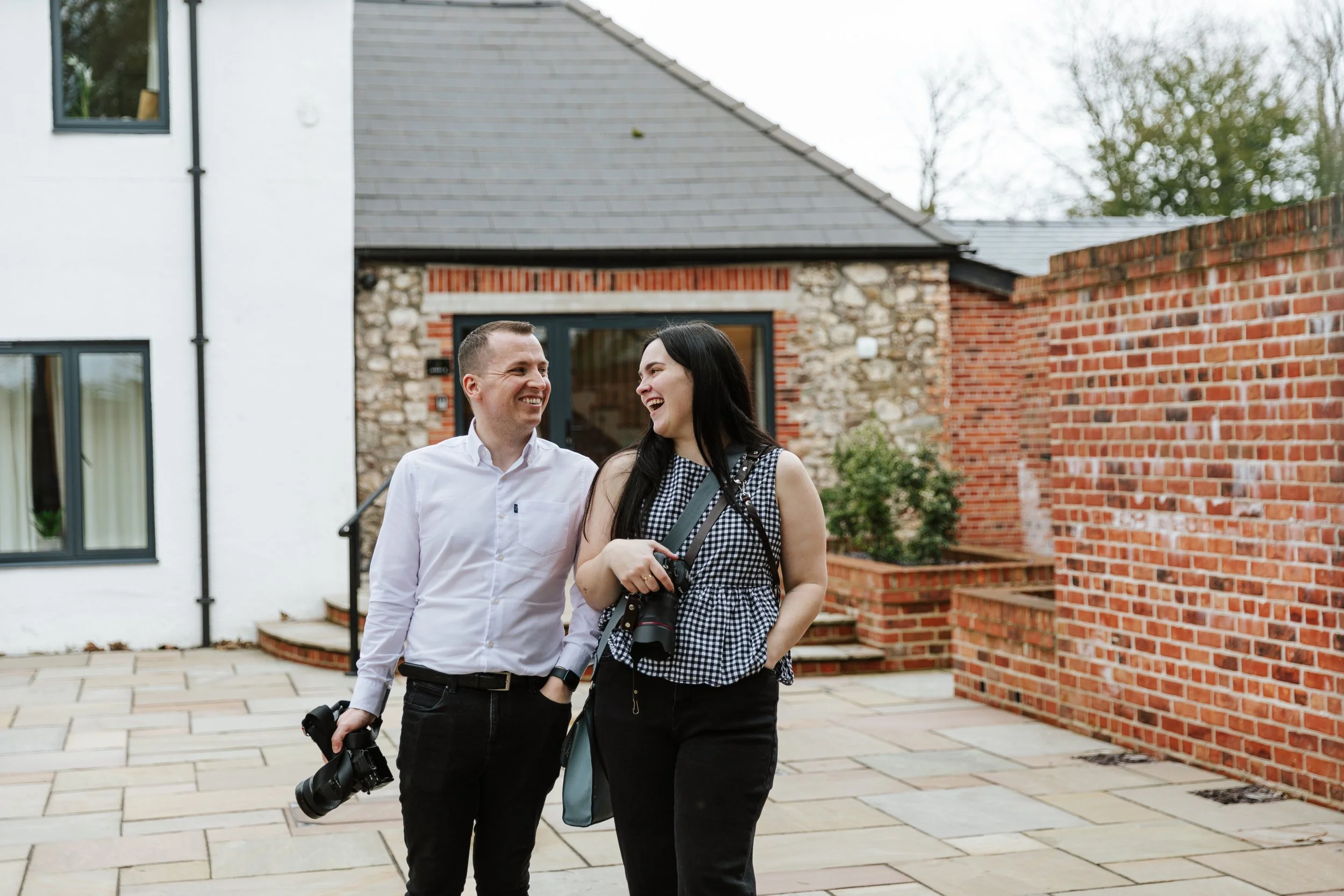 A man and woman are walking and talking in a residential backyard, smiling at each other. The woman is holding a camera, and the man has another camera in his hand. They are in front of a modern house with a white wall, large windows, and a brick and stone facade. There is a brick wall on the right and trees in the background.