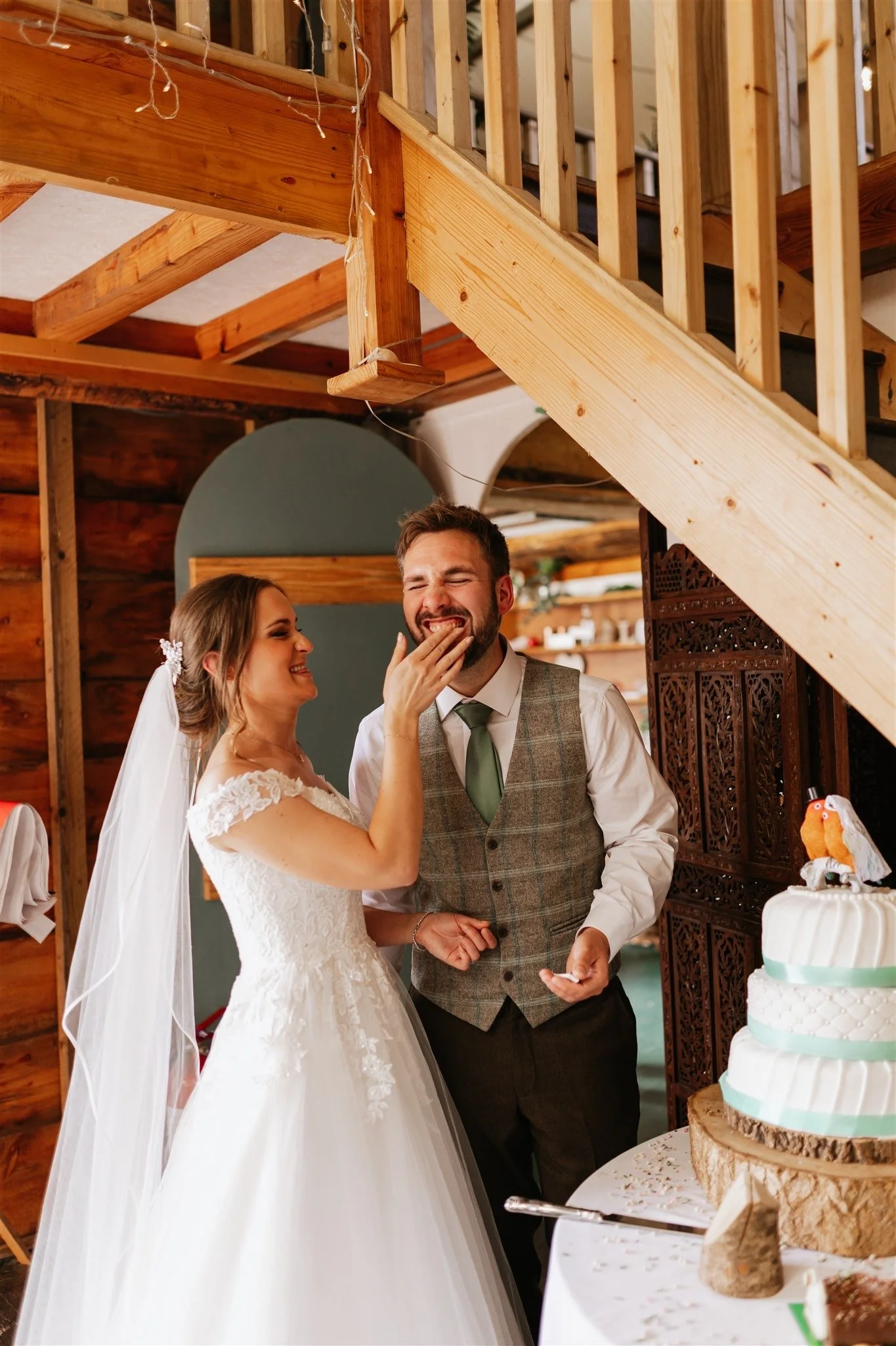 A bride and groom at their wedding reception standing near a wedding cake, with the bride smiling and the groom closing his eyes as she touches his face.