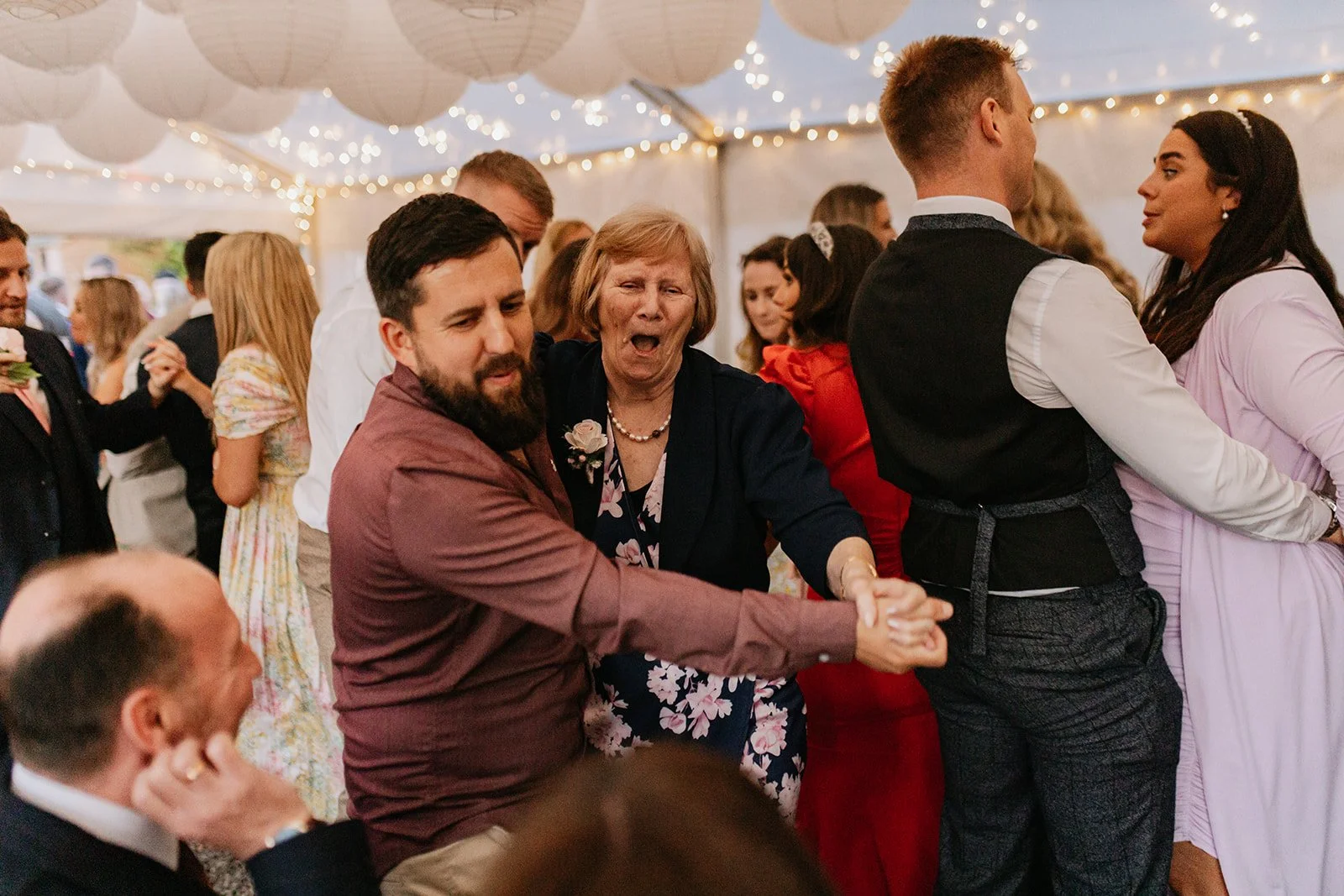 People dancing and celebrating at a wedding reception under a decorated tent with string lights and paper lanterns.