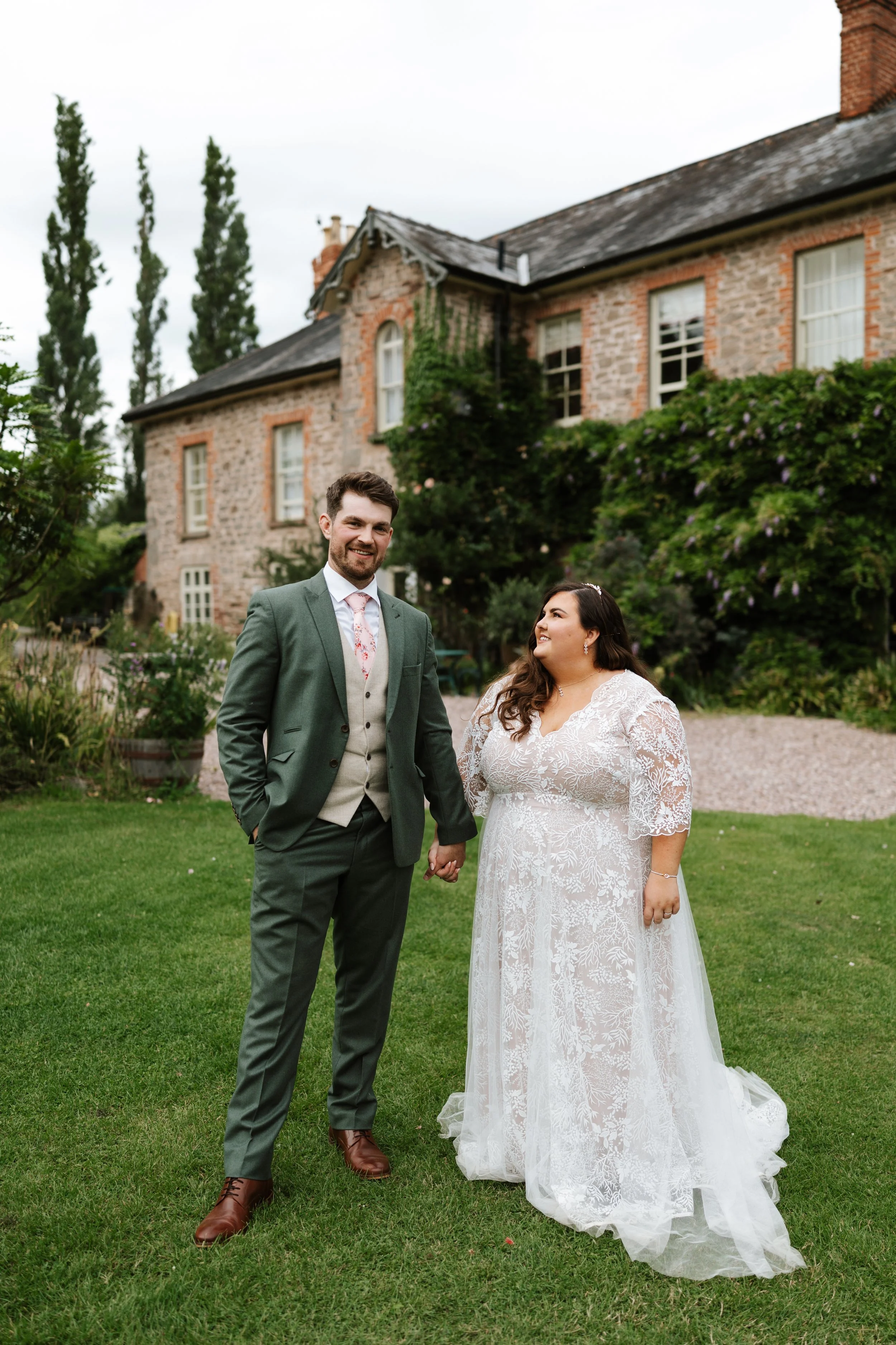 A man and woman in wedding attire holding hands in front of a stone house with greenery outside.