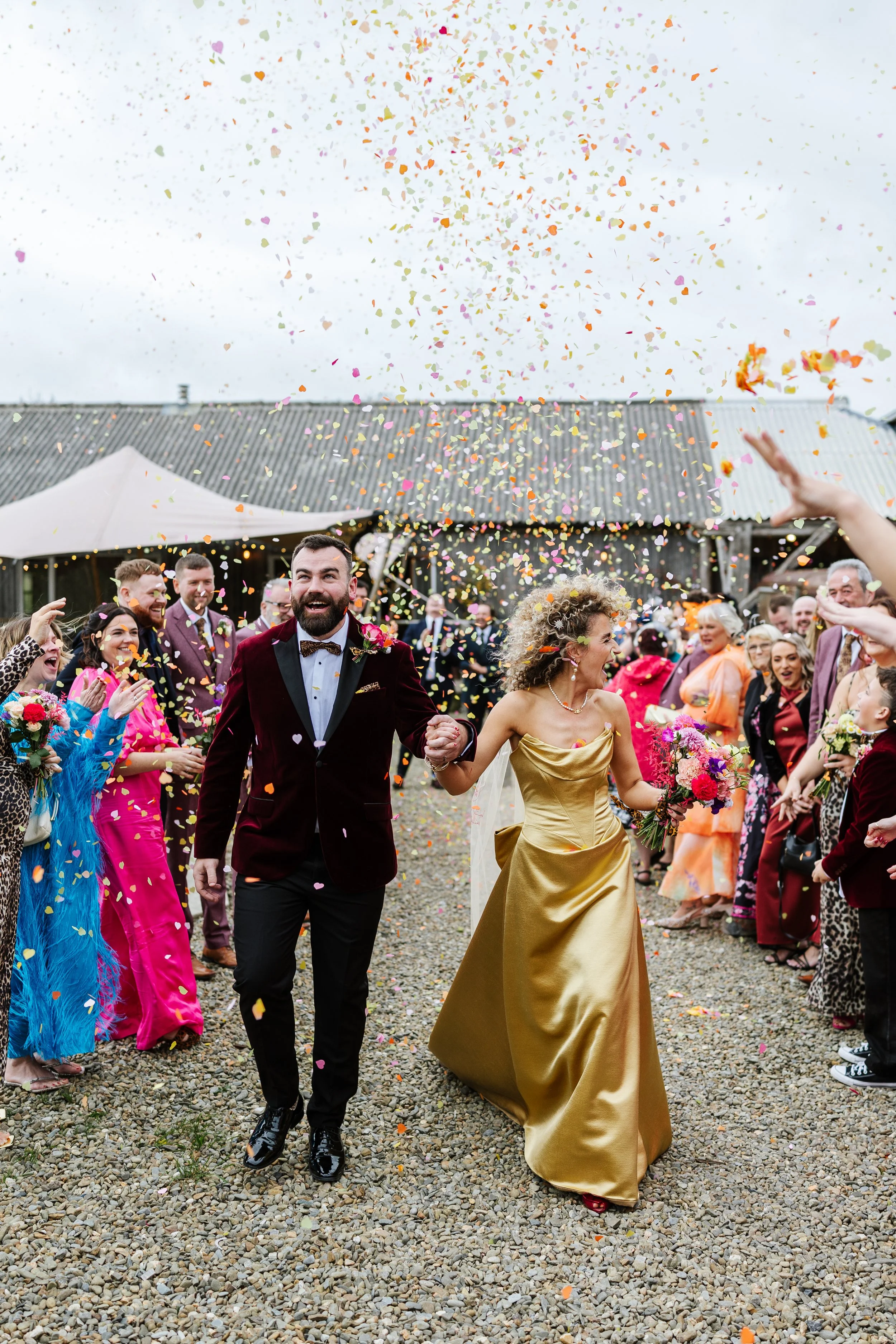 A wedding celebration with guests throwing colorful confetti as a bride and groom walk hand in hand outdoors.