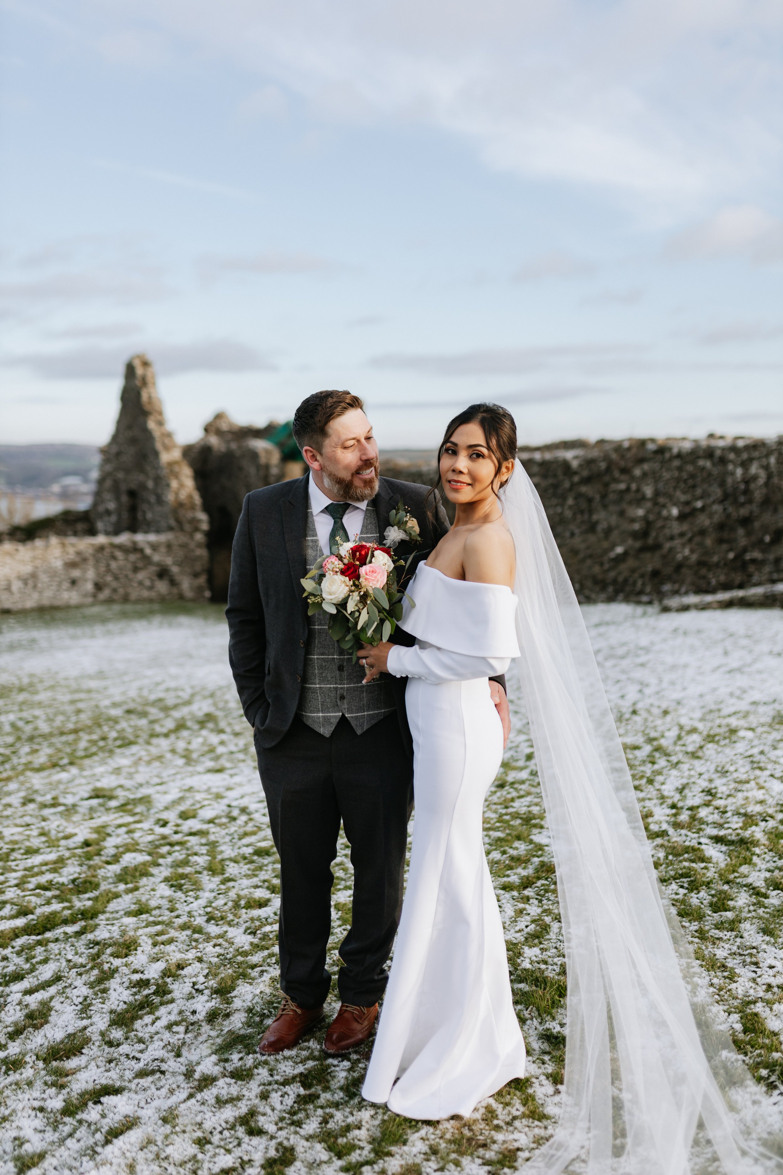 A bride and groom standing outdoors on a snowy field with ruins in the background, dressed in wedding attire, holding a bouquet of flowers.