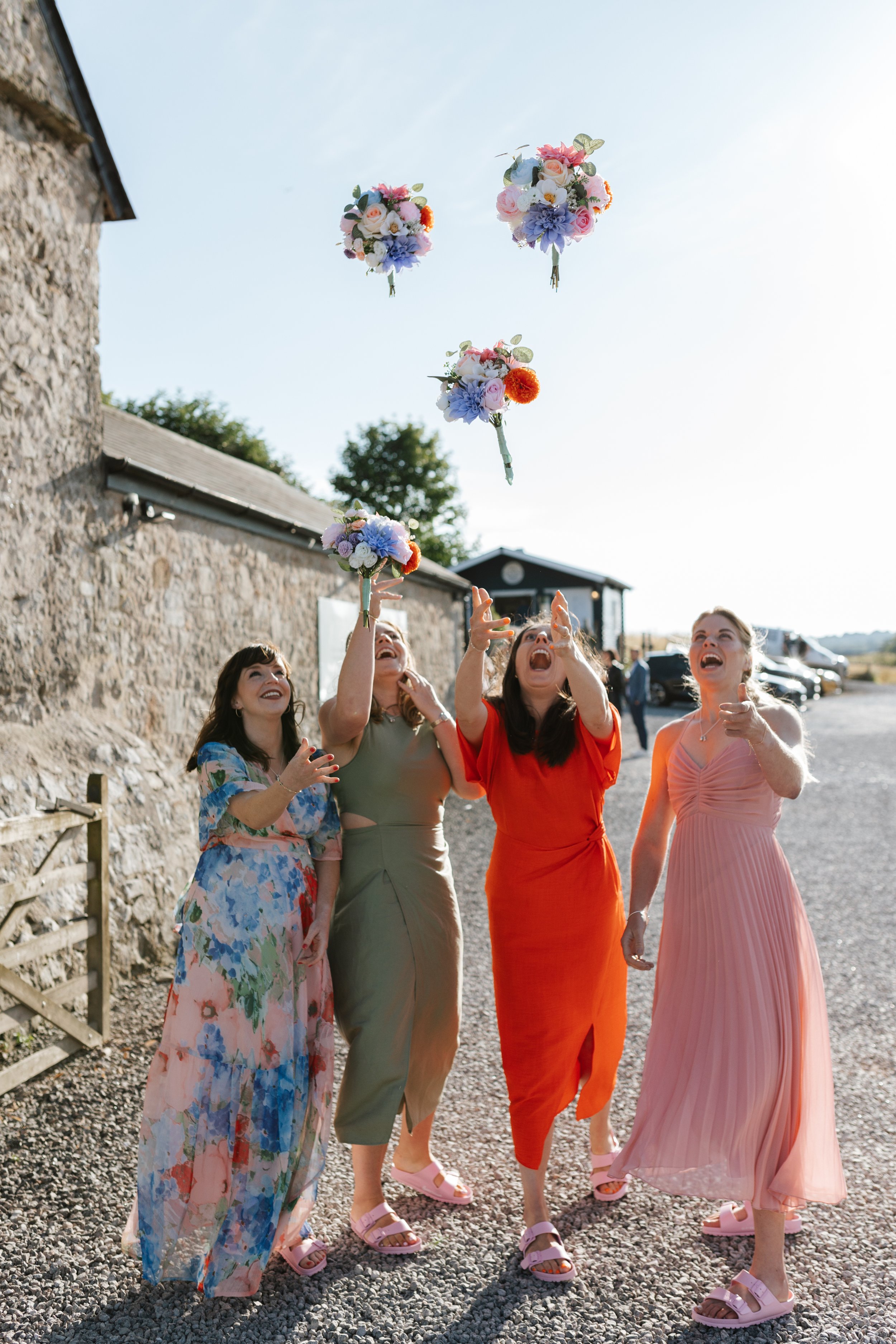Five women wearing colorful dresses are outdoors, tossing bouquets of flowers into the air during a celebration or wedding.