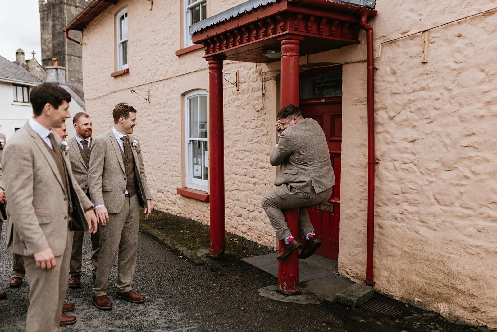 A man in a gray suit is sitting on a red porch post, holding his head, in front of a group of four men in suits, standing outside on a street with a beige brick building. The man appears to be joking or pretending to cry.