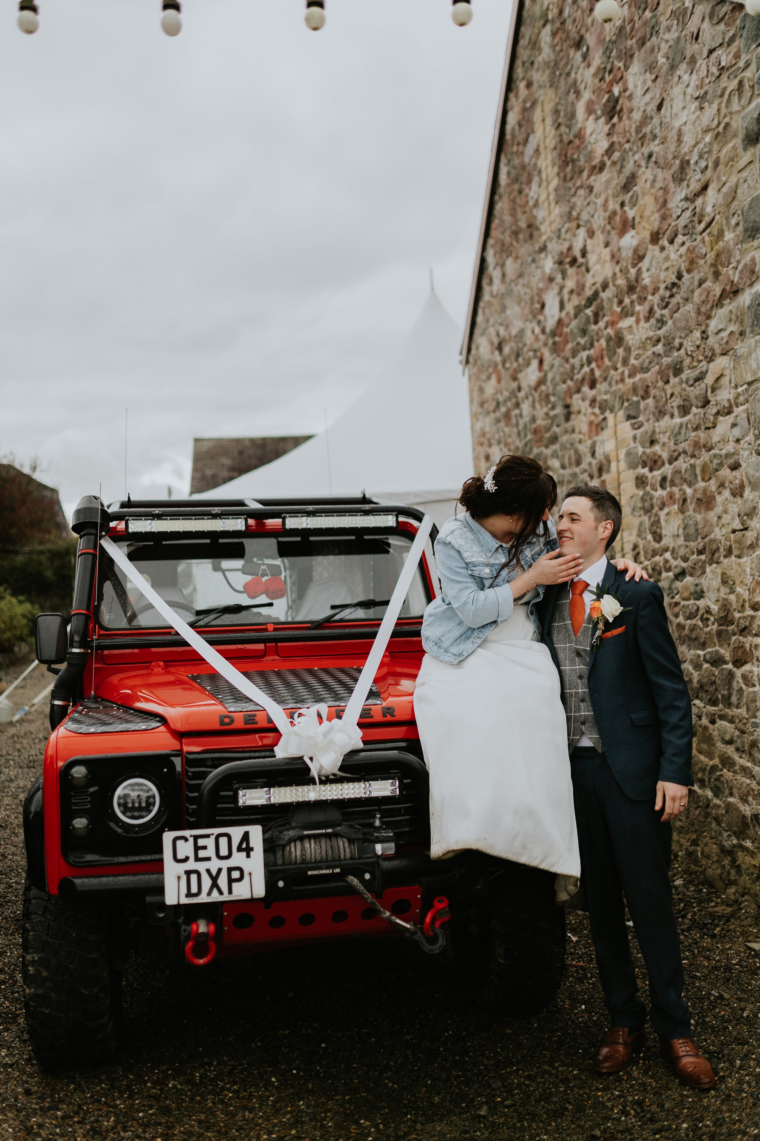 A couple dressed in wedding attire standing in front of a red off-road vehicle decorated with wedding ribbons and bows. The bride is sitting on the groom's lap, and they are smiling at each other. They are standing outdoors, near a stone building und