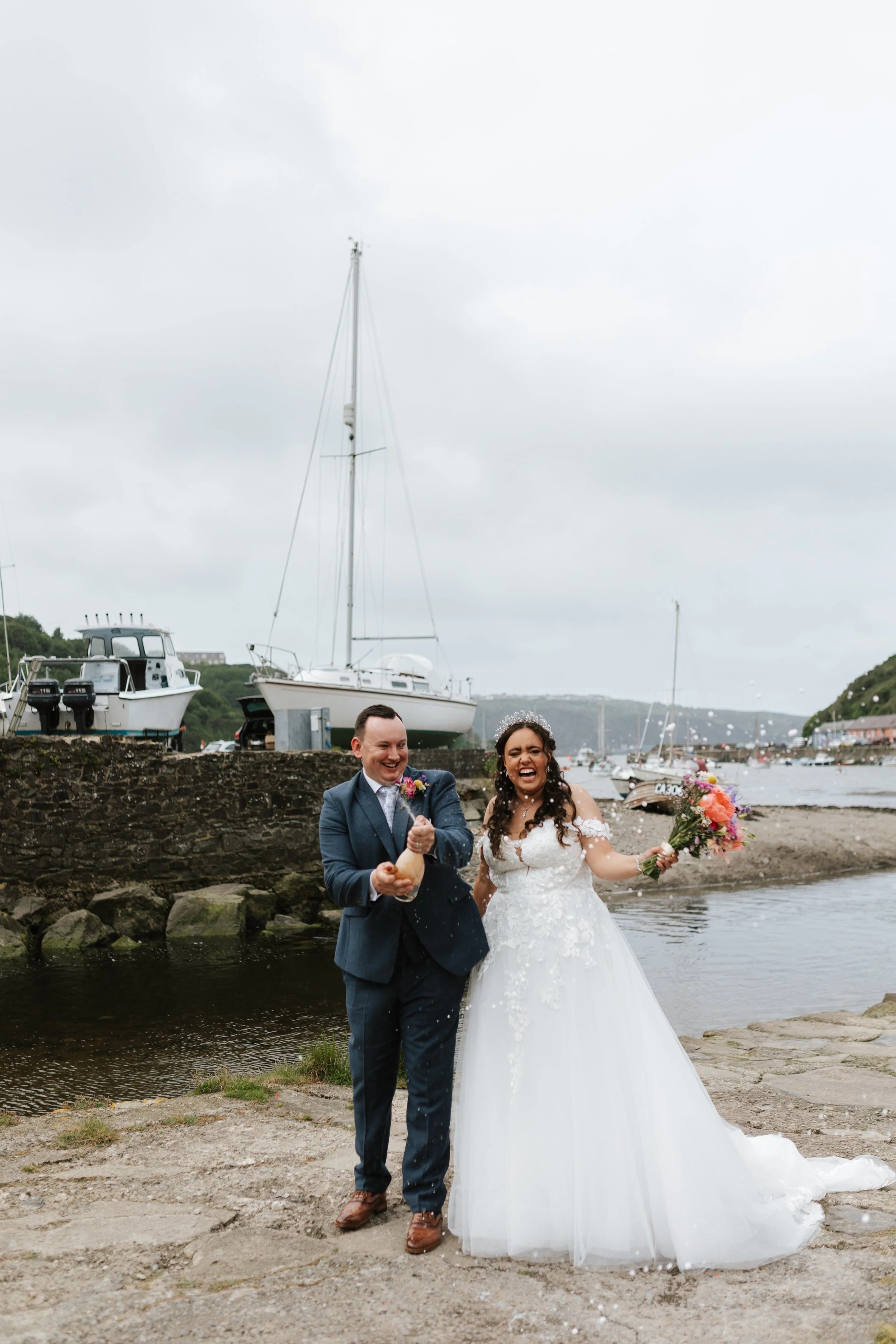 A bride and groom celebrating their wedding near a harbor, with boats and cloudy sky in the background. The bride is holding a bouquet and smiling, while the groom is dressed in a suit, holding champagne, both standing by the water.