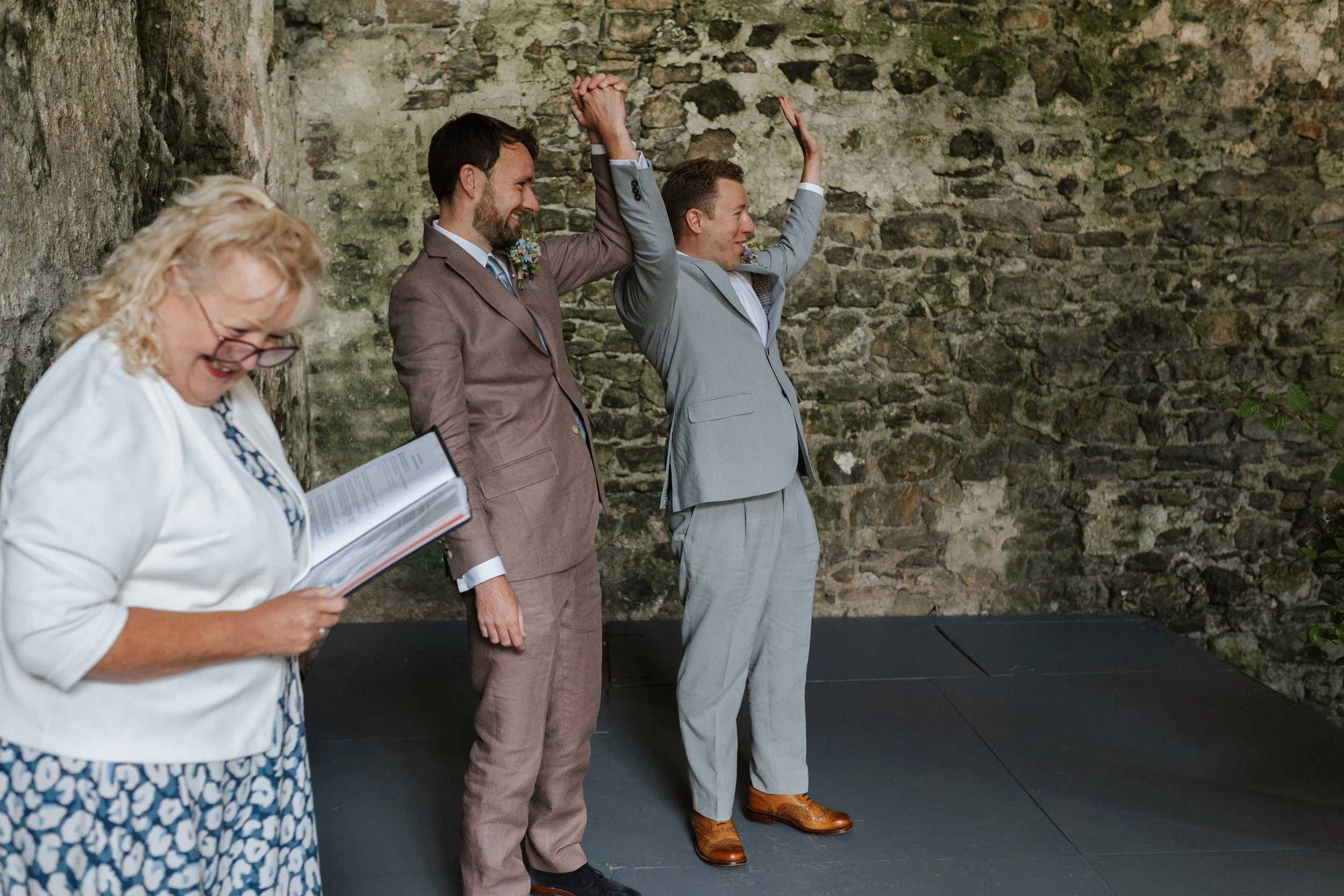 A group of people participating in a wedding ceremony outdoors, standing against a stone wall, with two men in suits raising their hands in a celebratory gesture, and a woman reading from a book or program.