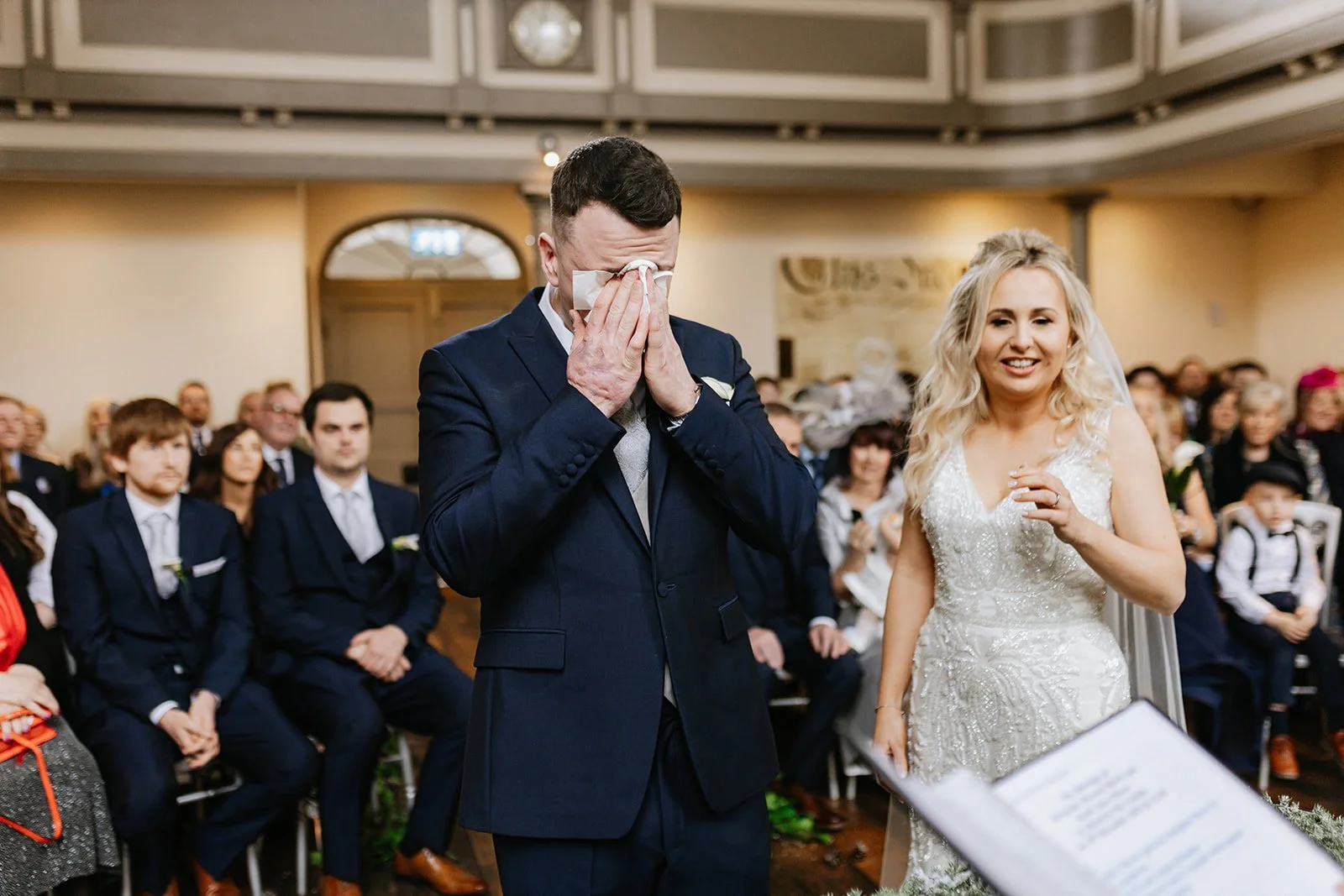 A wedding ceremony with a man in a navy suit crying and wiping tears with a tissue while a woman in a white wedding dress smiles beside him, surrounded by seated guests.