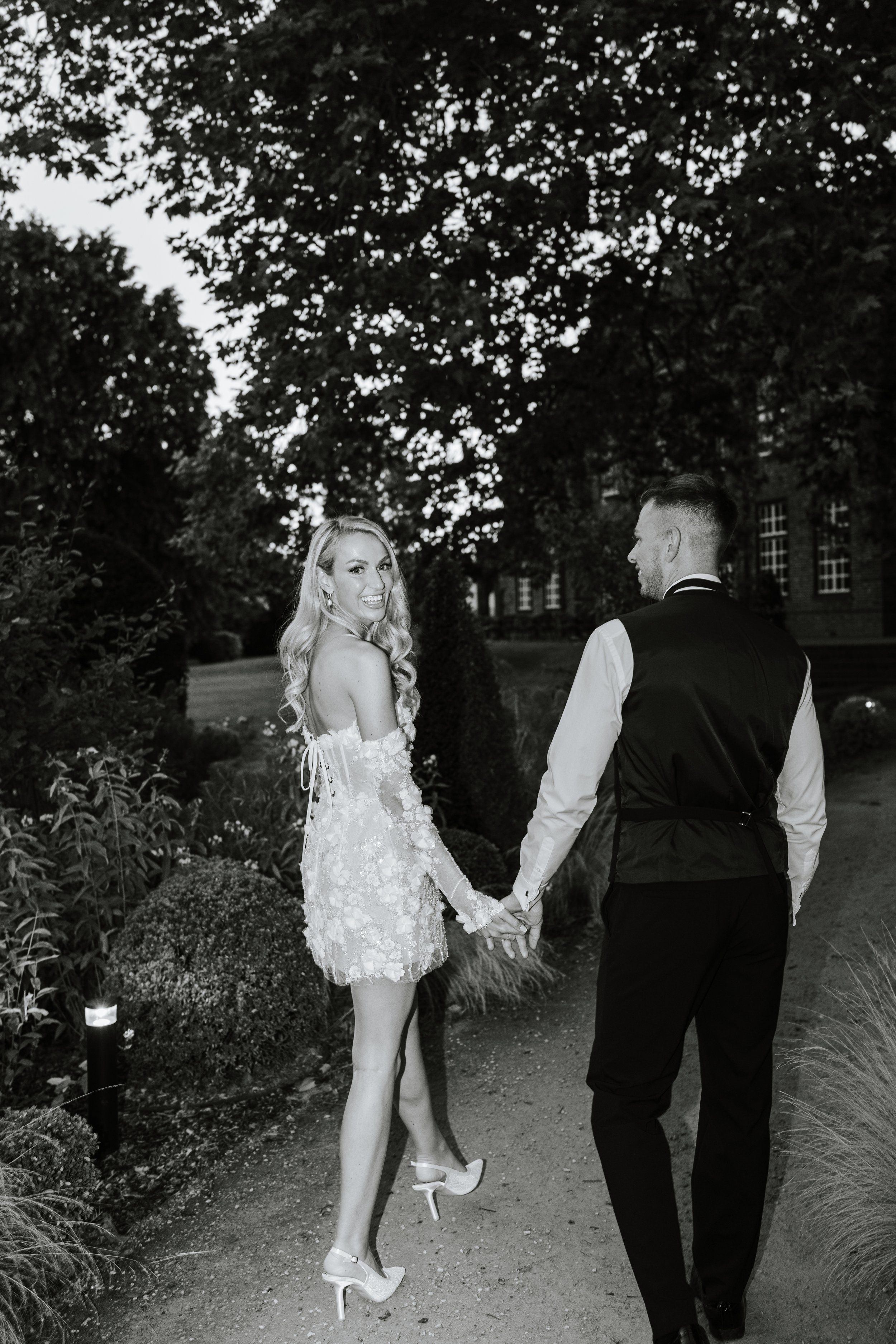 A black and white photo of a wedding couple holding hands outdoors; the woman is smiling and wearing a lace wedding dress and high heels, and the man is dressed in a vest and shirt, facing her with a tree and building in the background.
