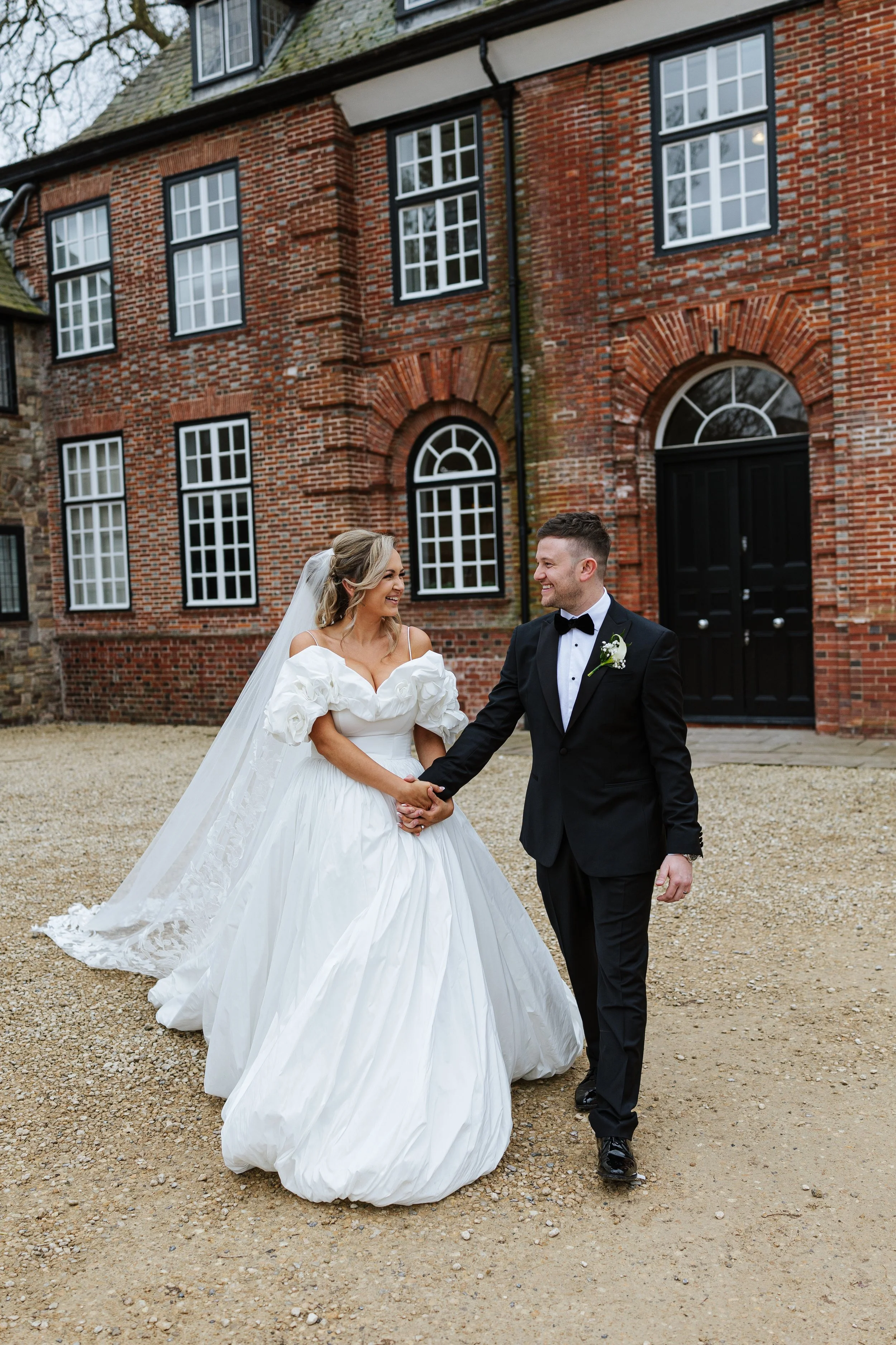 A bride and groom holding hands outside a brick building during their wedding, smiling at each other.