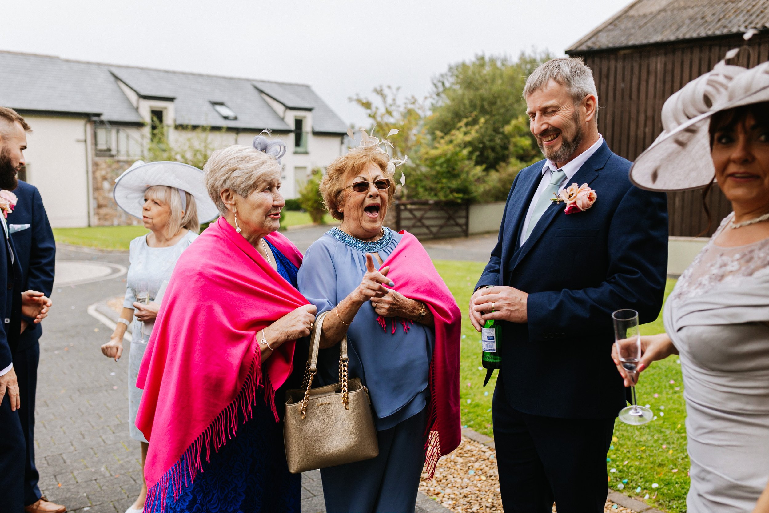 A group of people at an outdoor celebration, including elderly women with decorative hats and a man in a suit smiling, engaging in conversation and holding drinks.