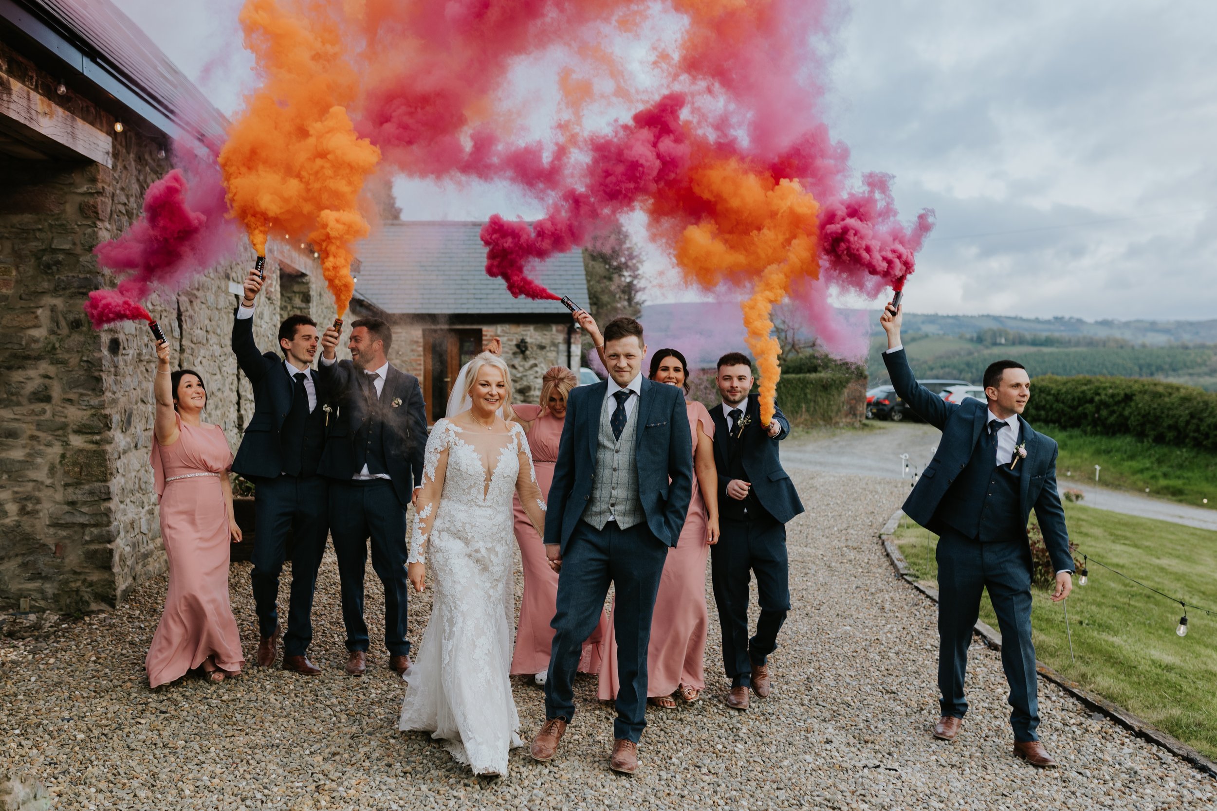 A group of people, including a bride in a white wedding dress, walking outdoors on a gravel path with smiles. They are holding colorful smoke grenades emitting pink and orange smoke, possibly celebrating a wedding or special event.