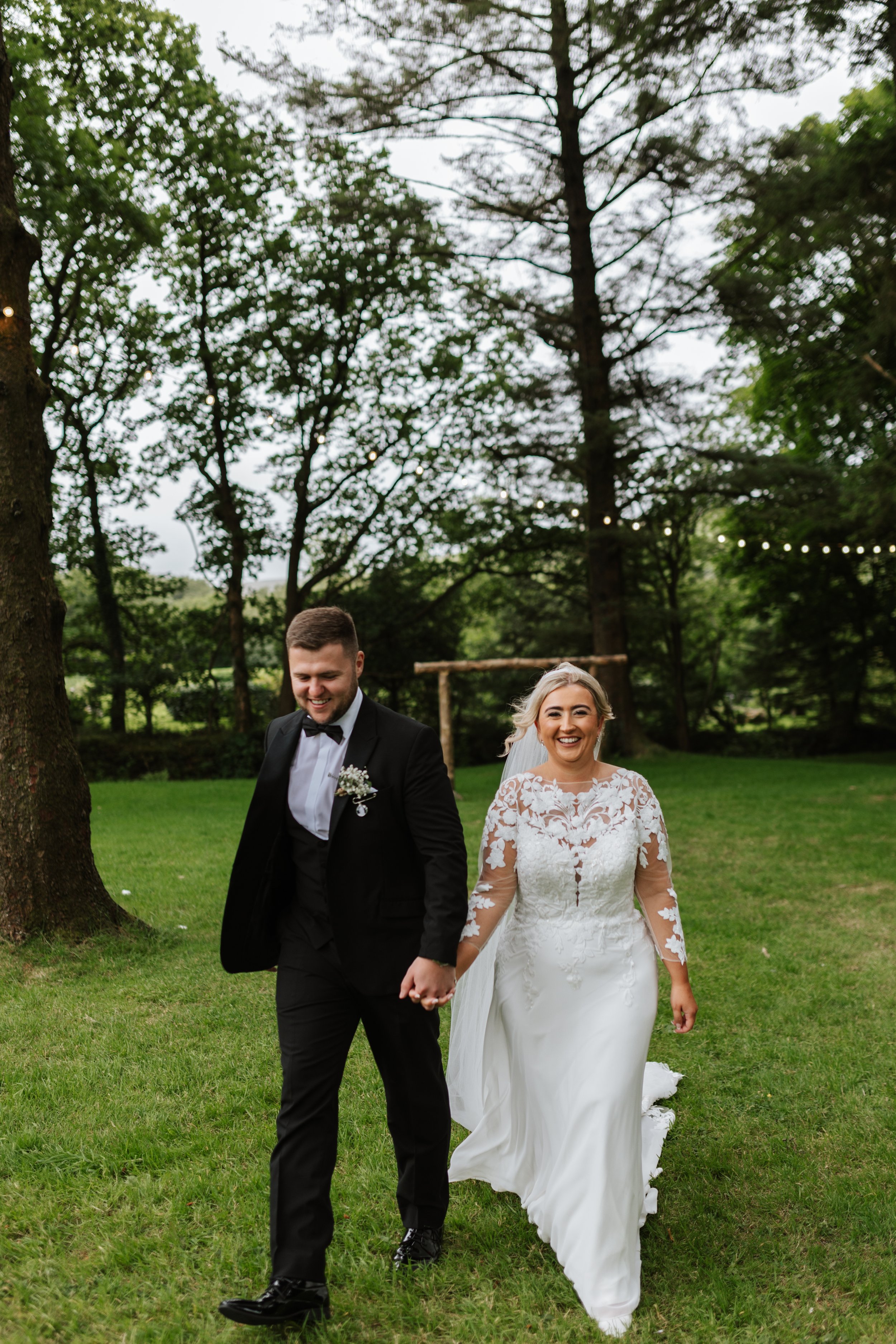 A smiling bride and groom walking hand in hand outdoors in a green park with trees and string lights overhead, during a wedding celebration.