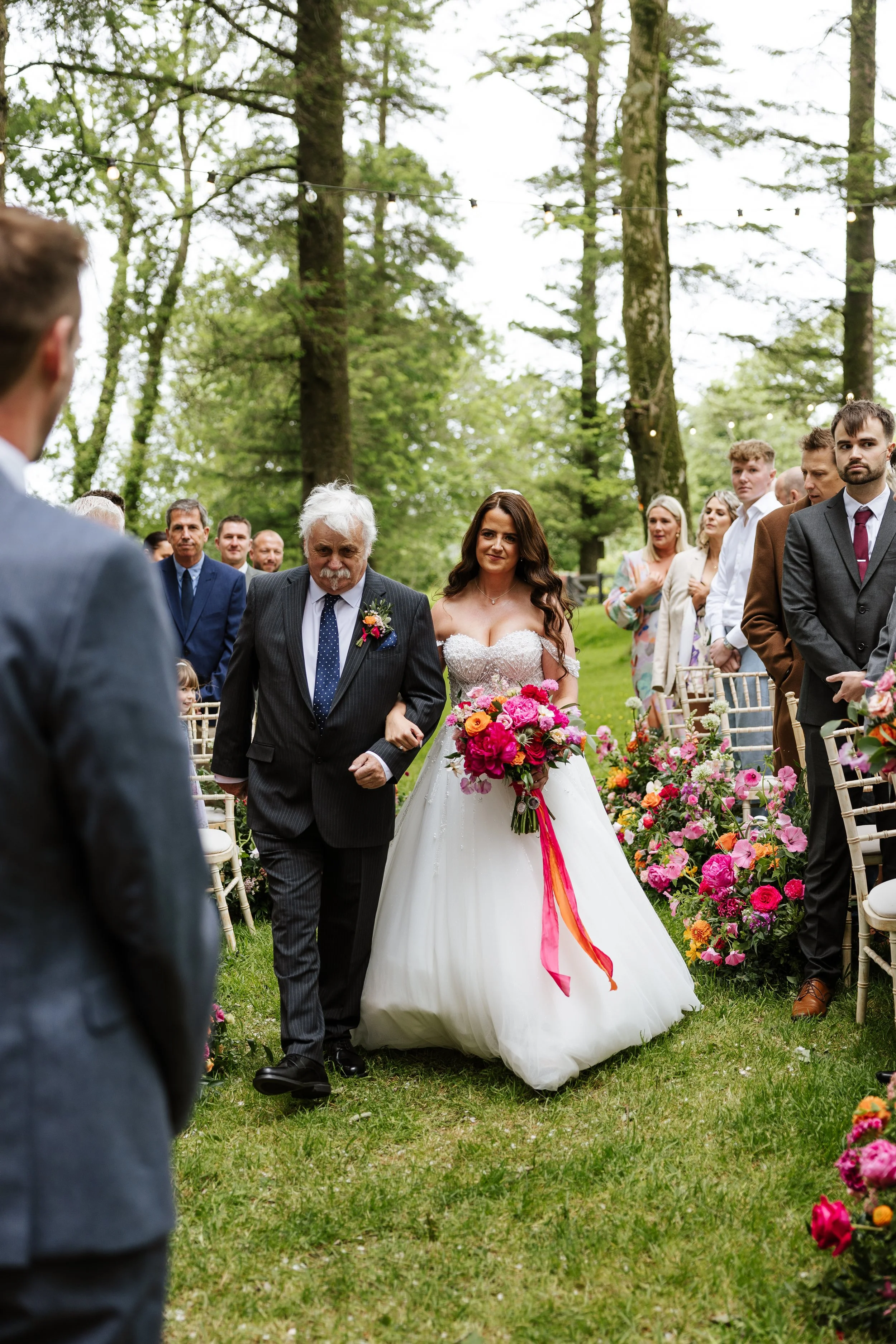 A bride in a white wedding dress holding a bouquet of pink, orange, and yellow flowers, walking down an aisle in a forested outdoor wedding ceremony, accompanied by a man in a black suit with a boutonniere, surrounded by wedding guests.