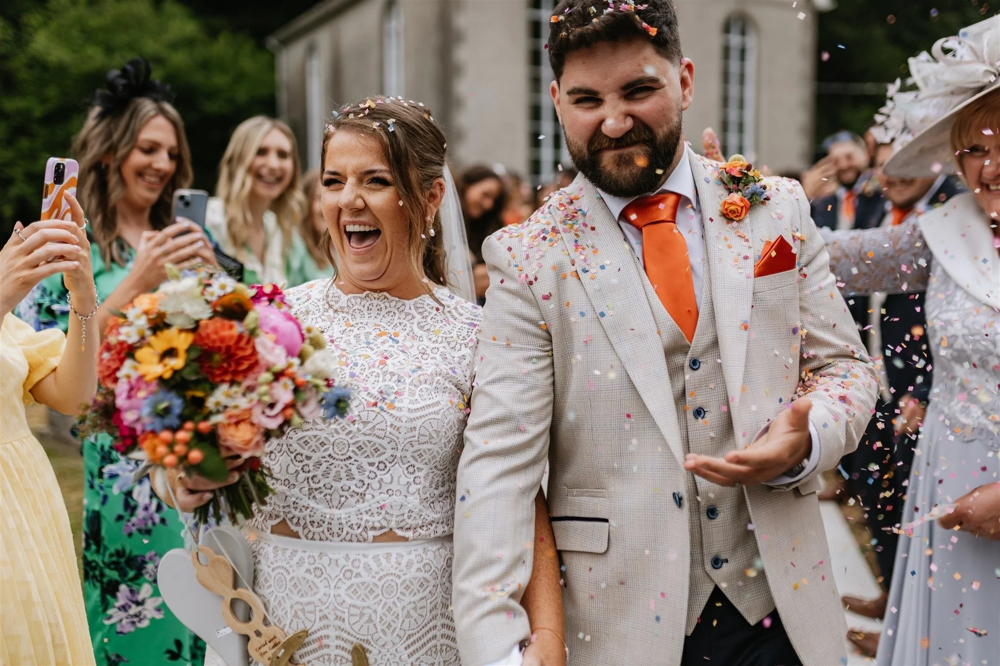 A bride and groom smiling and celebrating with guests at their wedding outside. The bride has a bouquet of colorful flowers and the groom is in a light-colored suit with an orange tie. Confetti is falling around them.