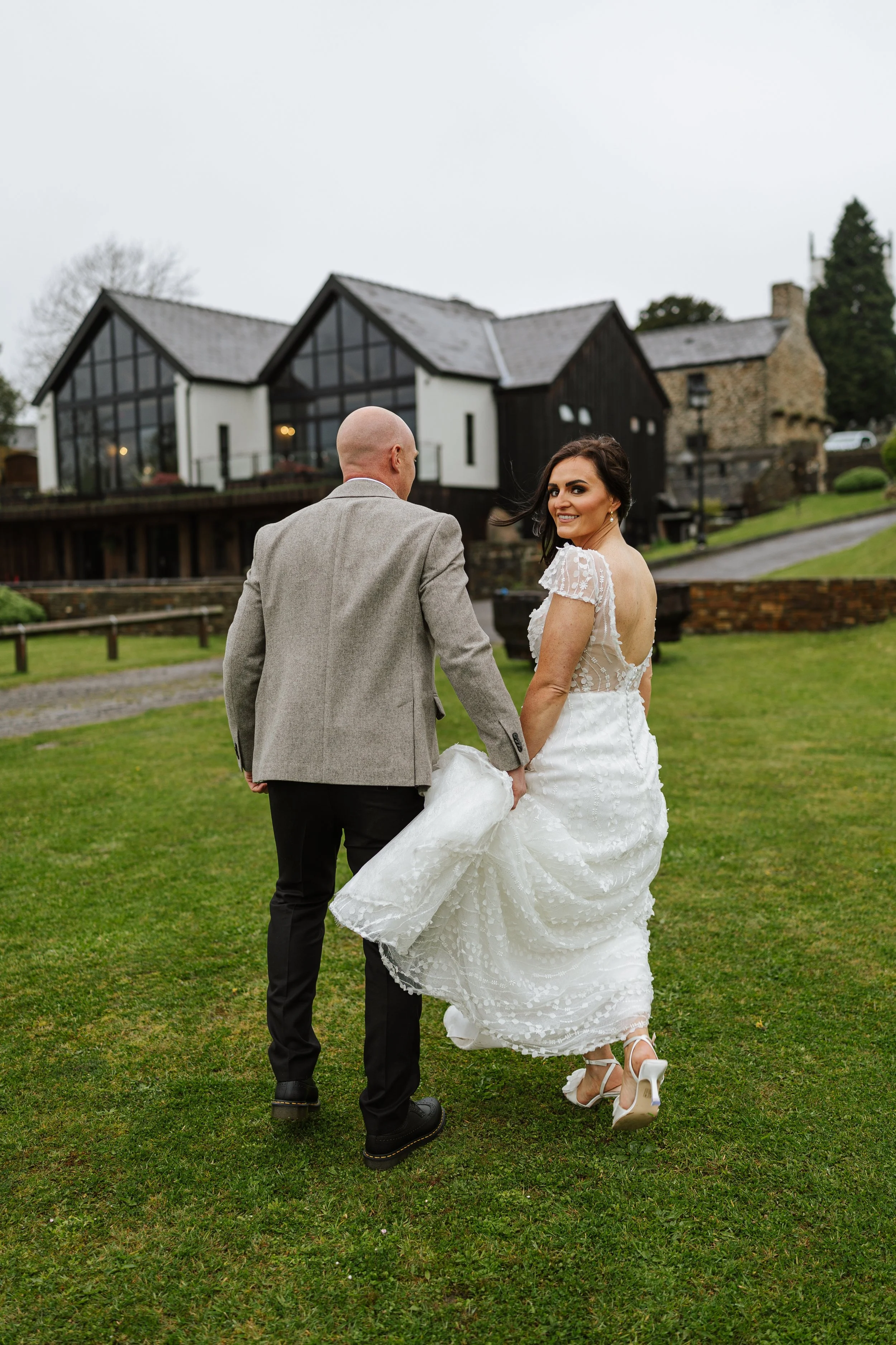 A bride and groom walking hand in hand on a grassy lawn outside a modern house on a cloudy day. The bride is smiling and looking back at the camera, wearing a white wedding dress and heels. The groom is wearing a gray suit, facing away from the camer