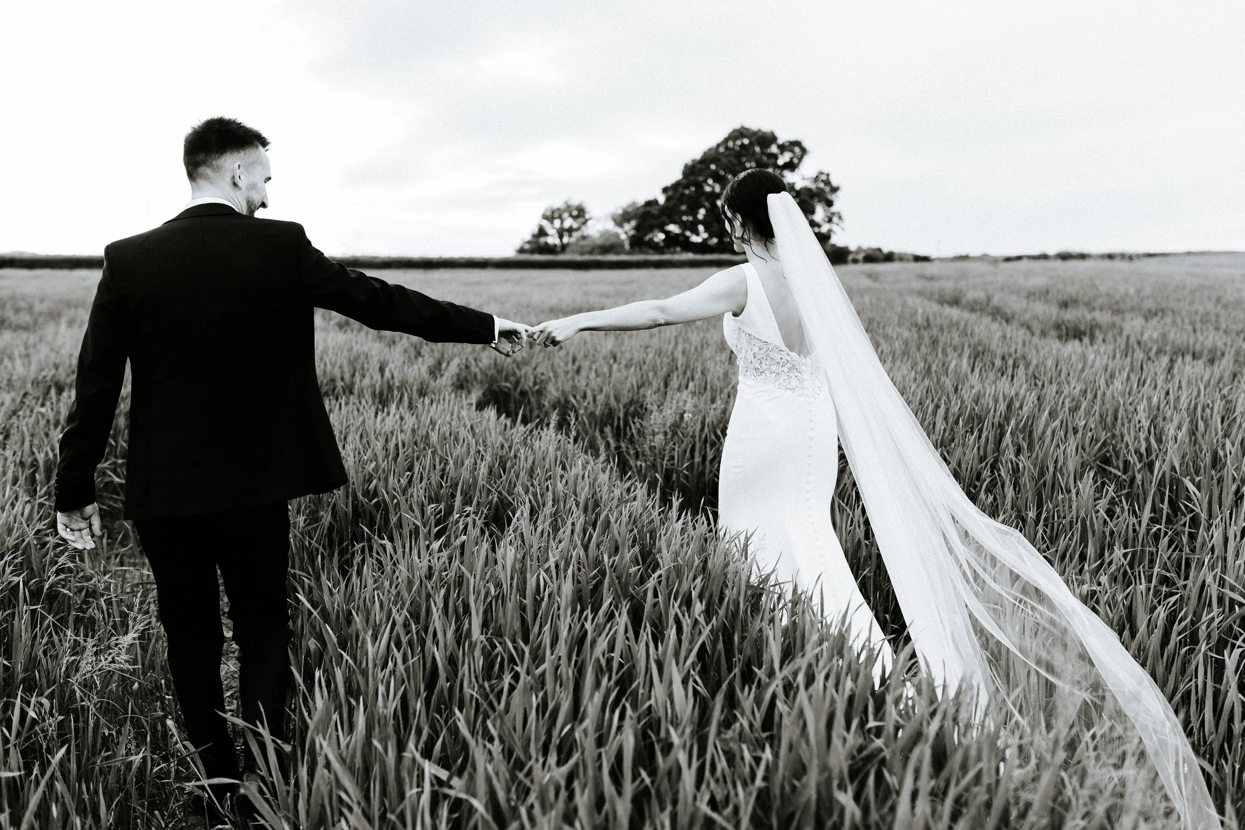 A black-and-white photo of a bride and groom holding hands in a field of tall grass, with trees and a cloudy sky in the background.