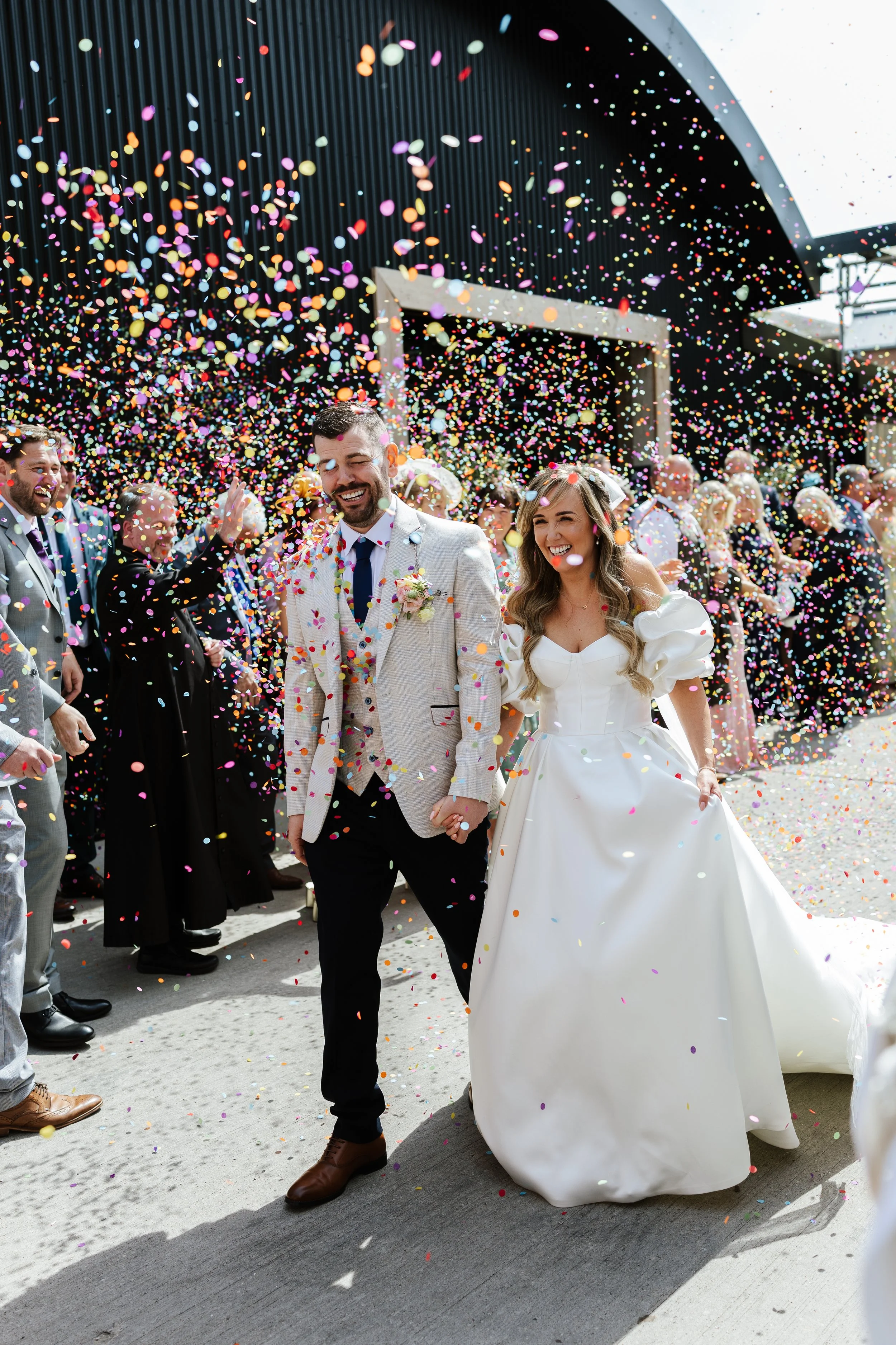 A newlywed couple holding hands smiling as confetti falls around them at their wedding celebration outdoors with friends and family in the background.
