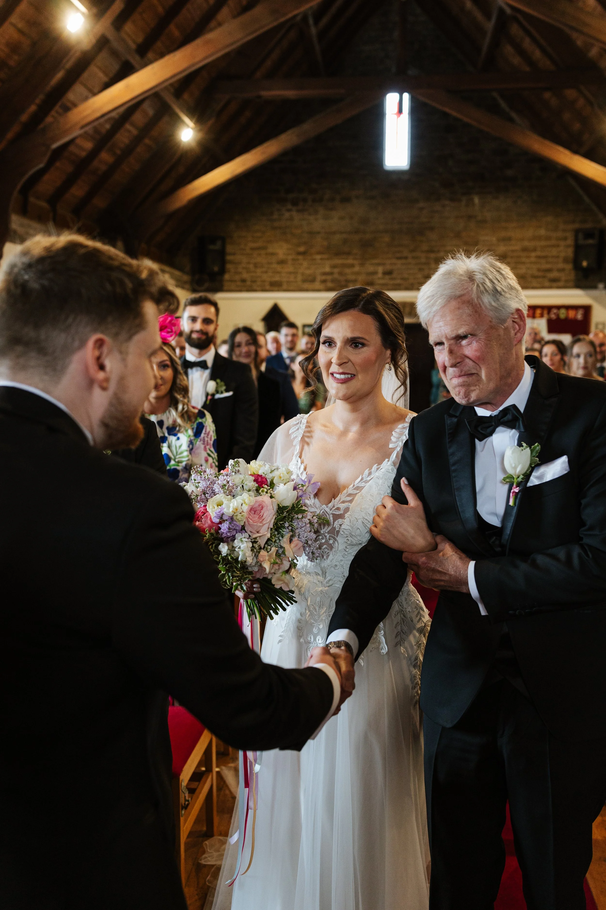 A bride and an older man, likely her father, holding hands during a wedding ceremony inside a church. The bride is holding a bouquet of flowers, and the scene is witnessed by a group of guests in formal attire.