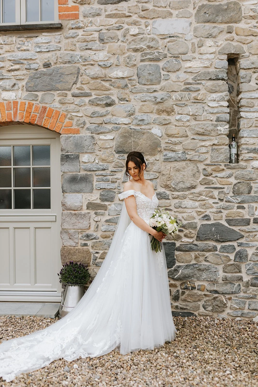 A bride in a white wedding dress holding a bouquet of white flowers, standing in front of a rustic stone wall with a small window.