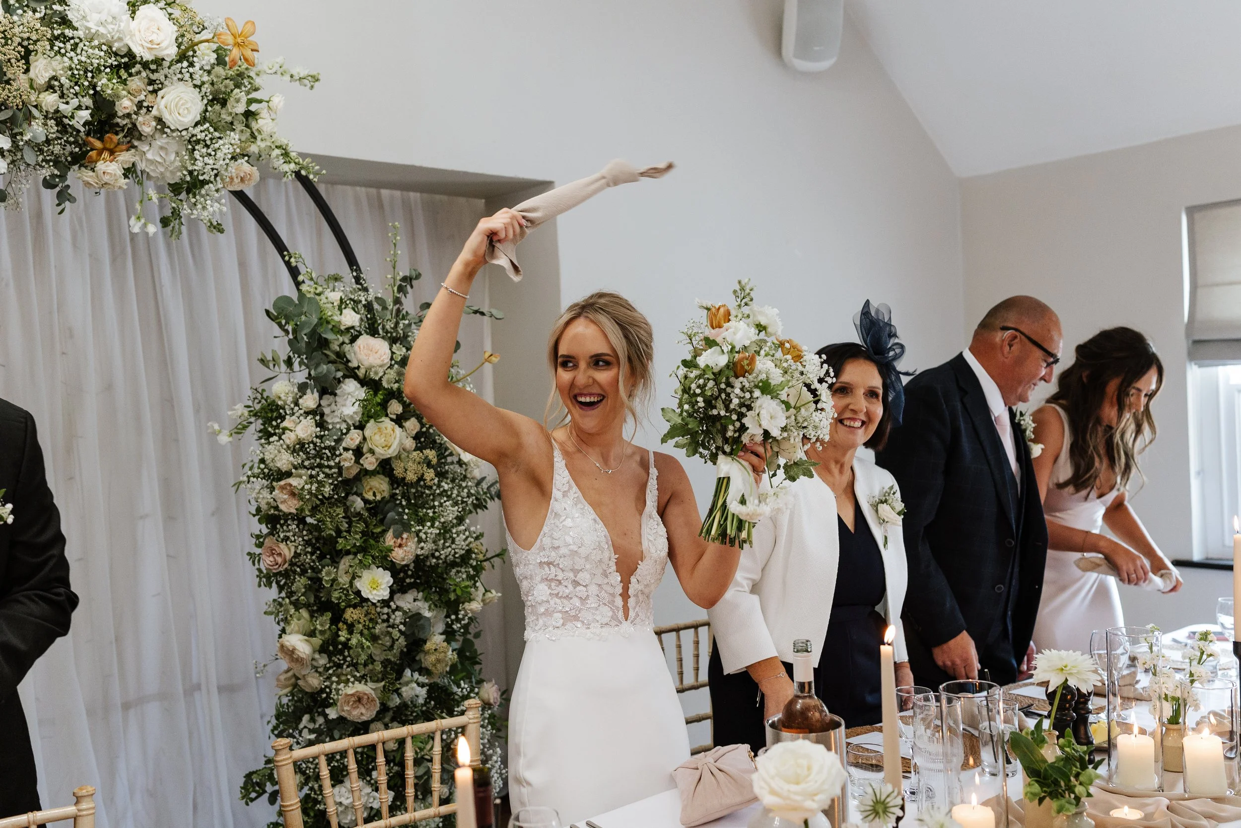 A bride in a white wedding gown holding a bouquet, standing at a wedding reception with other guests. There are floral arrangements and candles on the table in front of her.